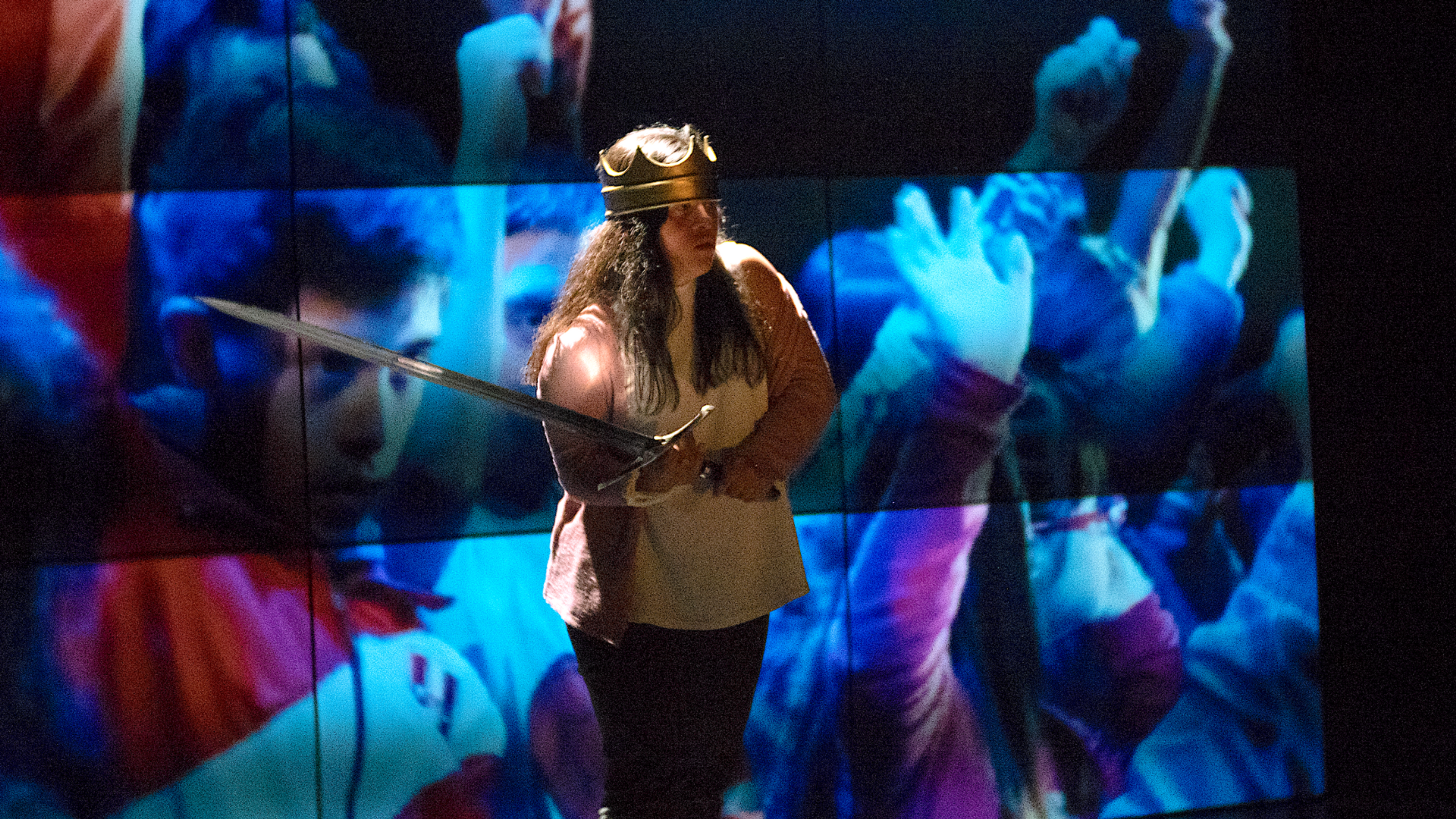 An actor wearing a crown and holding a sword stands in front of a background projection of people.