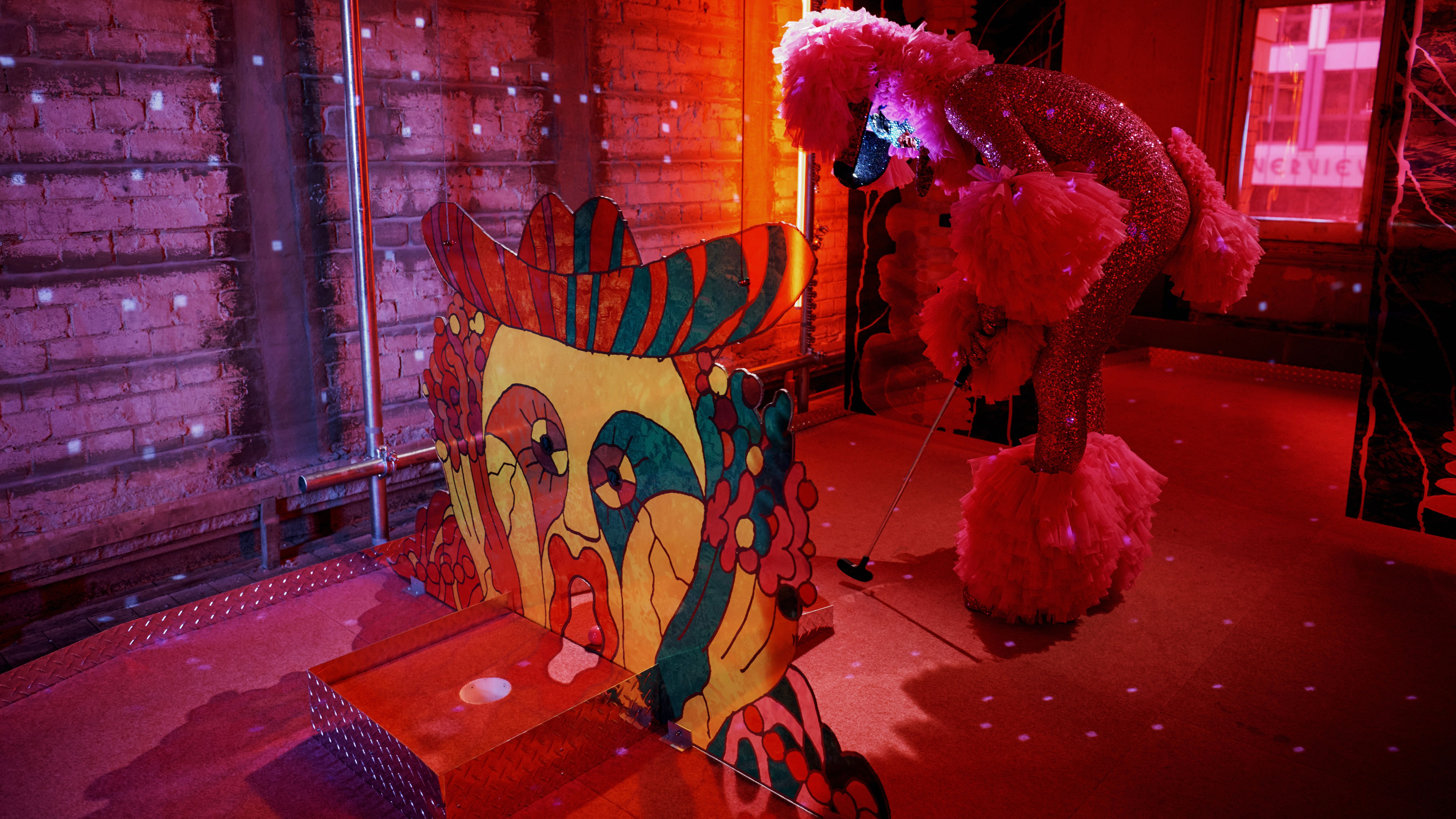 One of the Huxleys , dressed as a pink poodle, lining up to putt a ball through the mouth of an an abstracted sculpture of a face