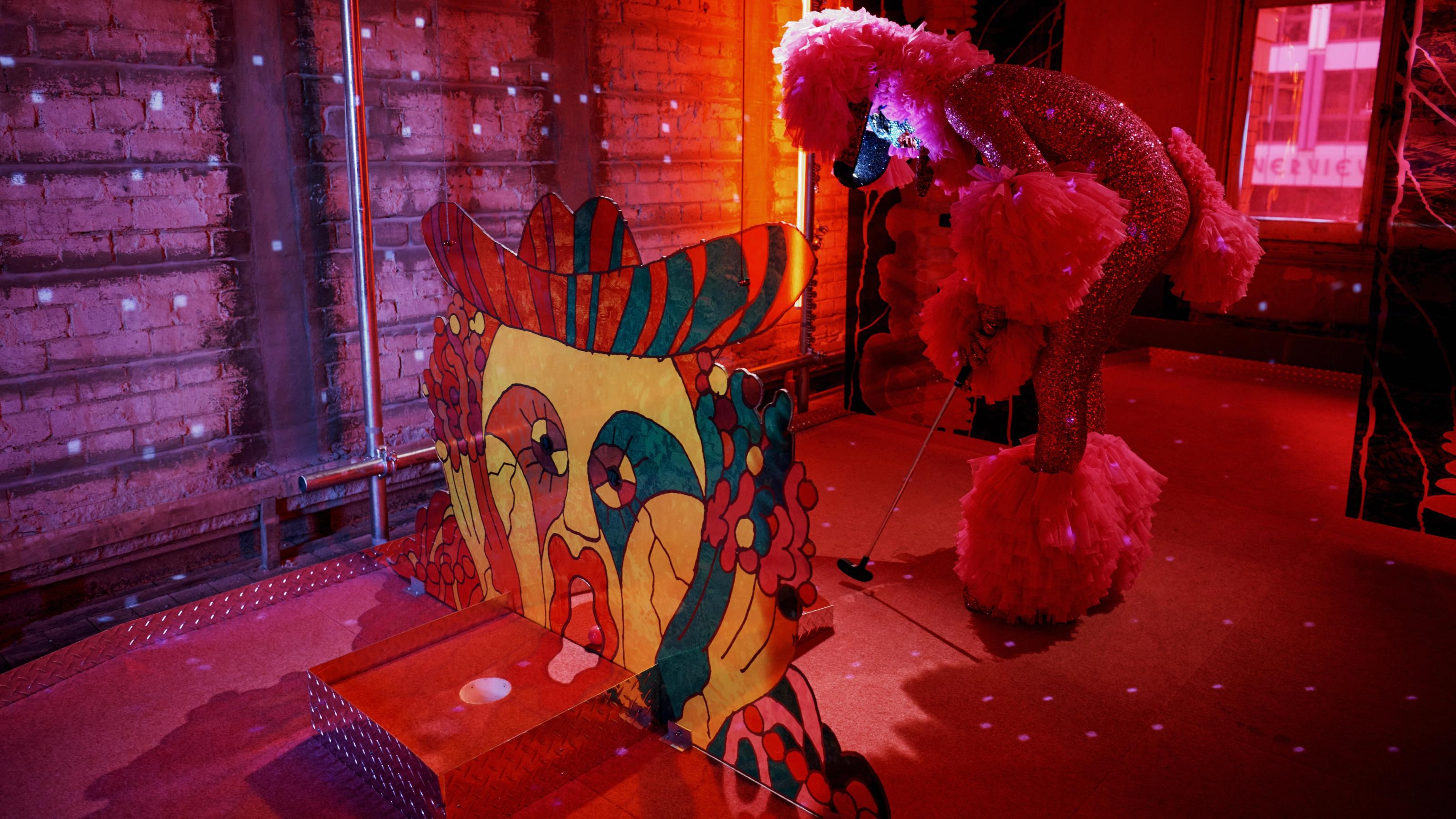 One of the Huxleys , dressed as a pink poodle, lining up to putt a ball through the mouth of an an abstracted sculpture of a face