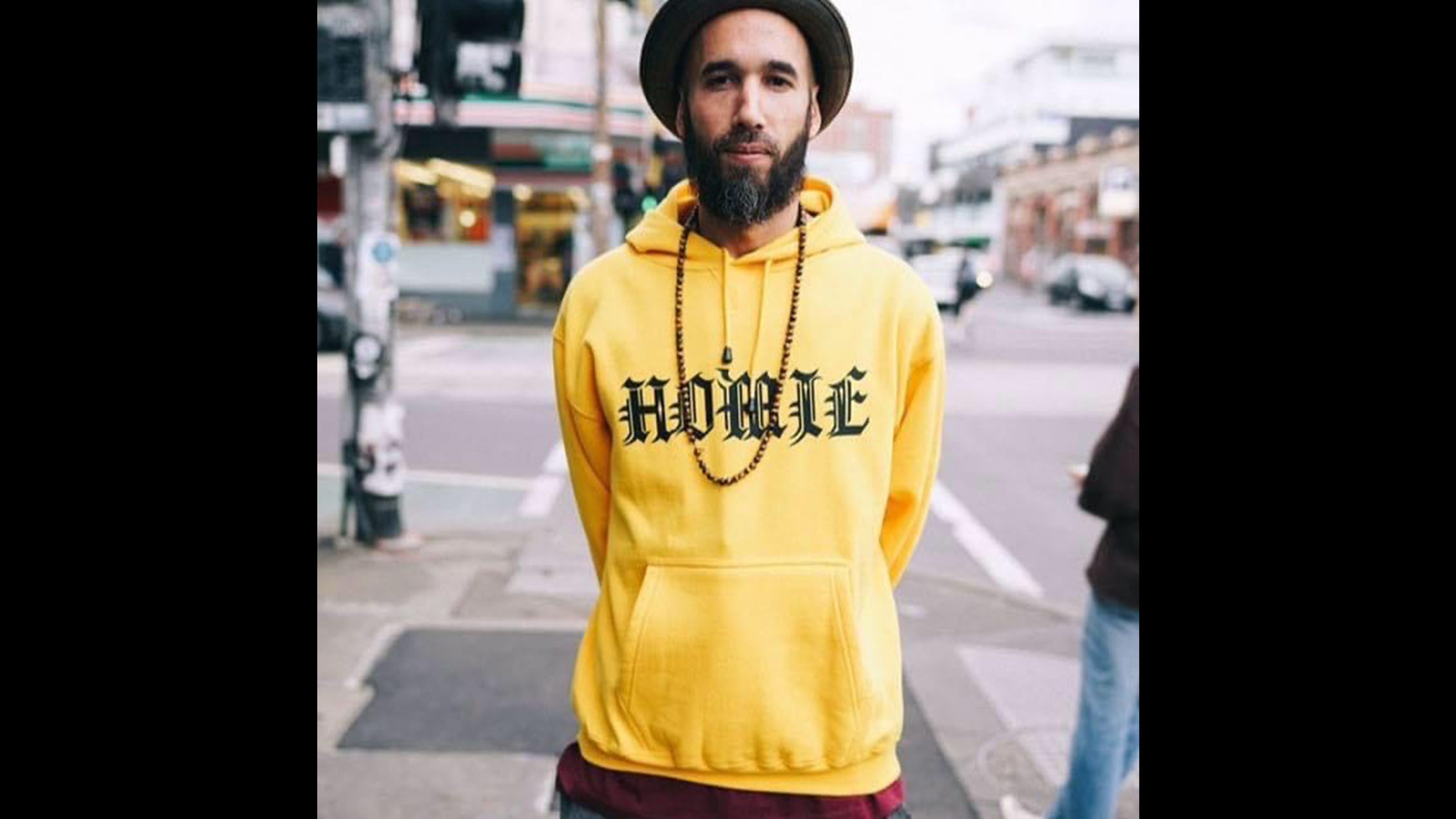DJ Sensiible J stands on a street facing the camera. He wears a hat and a long chain