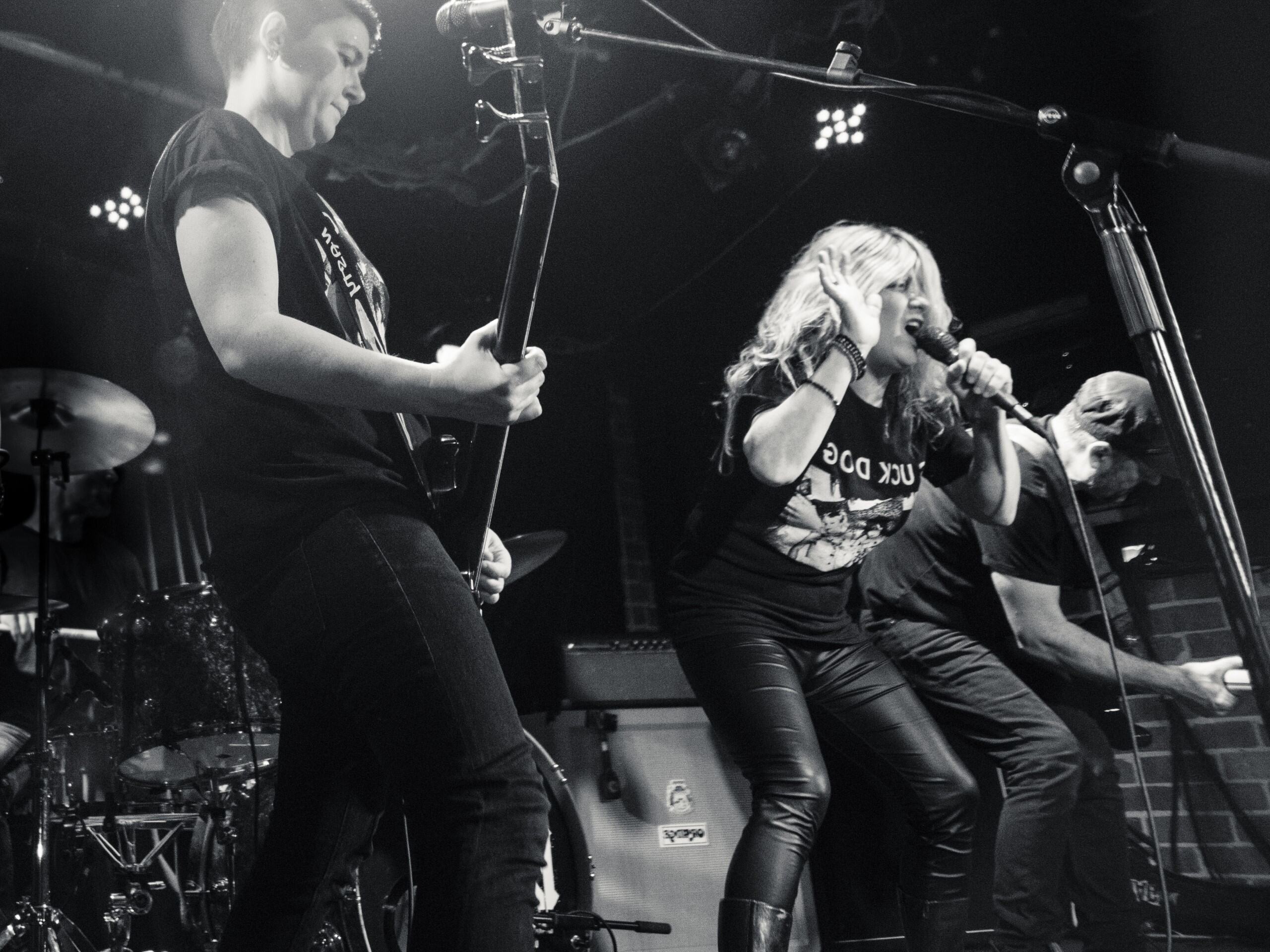 Three people are performing on stage in black and white. Two people on either side are playing guitar, while the person in the middle is singing.
