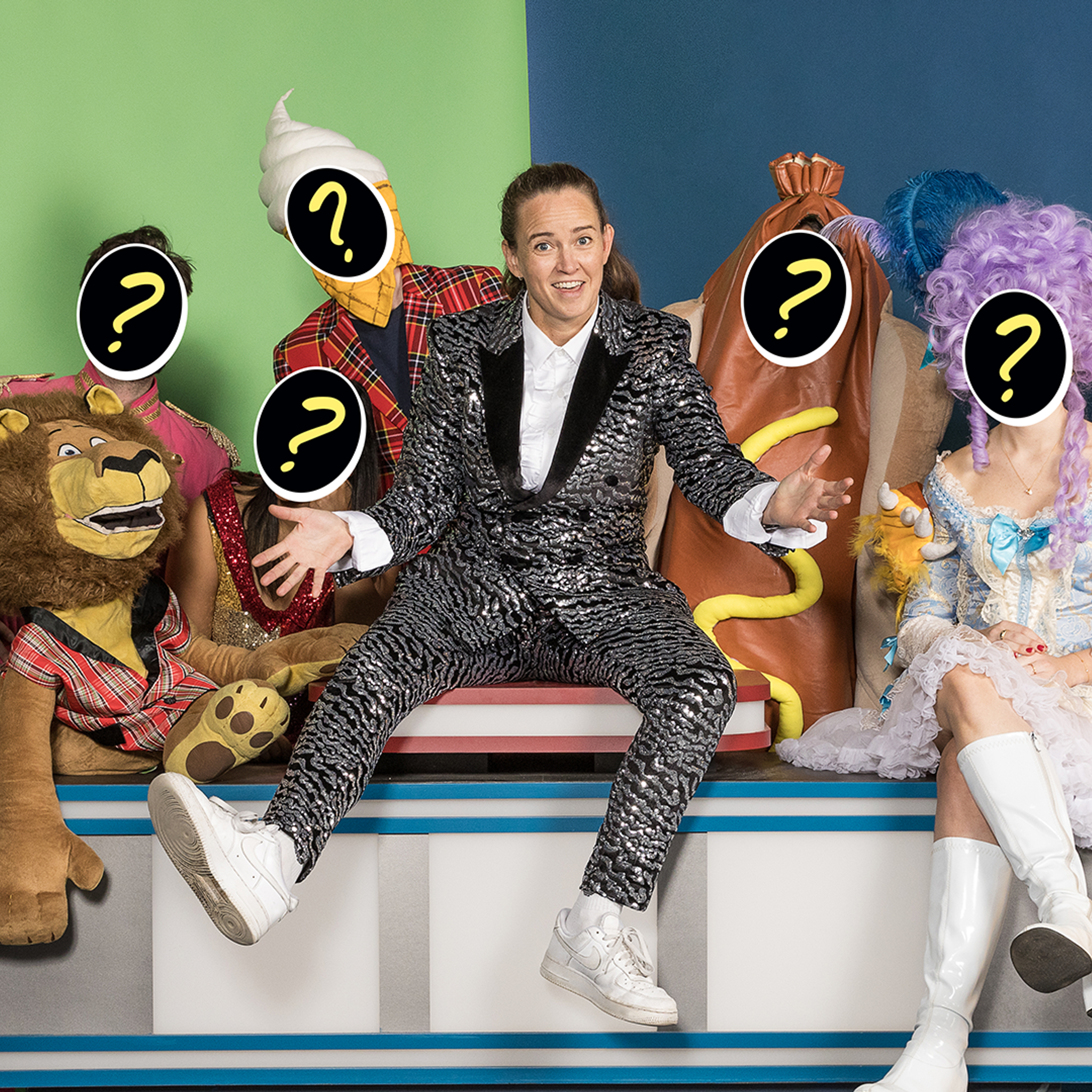 Zoë Coombs Marr dressed as a variety show host, surrounded by costumed characters with question marks superimposed on their faces