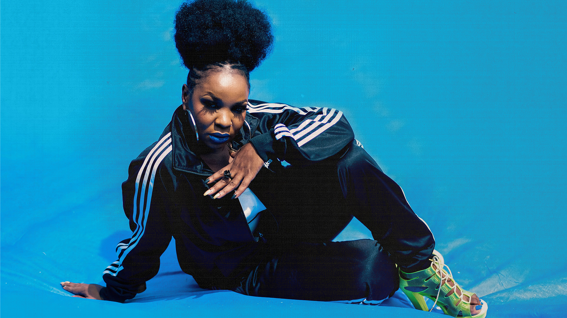 MC Yallah wears a full tracksuit and sits on the floor, leaning on their arm