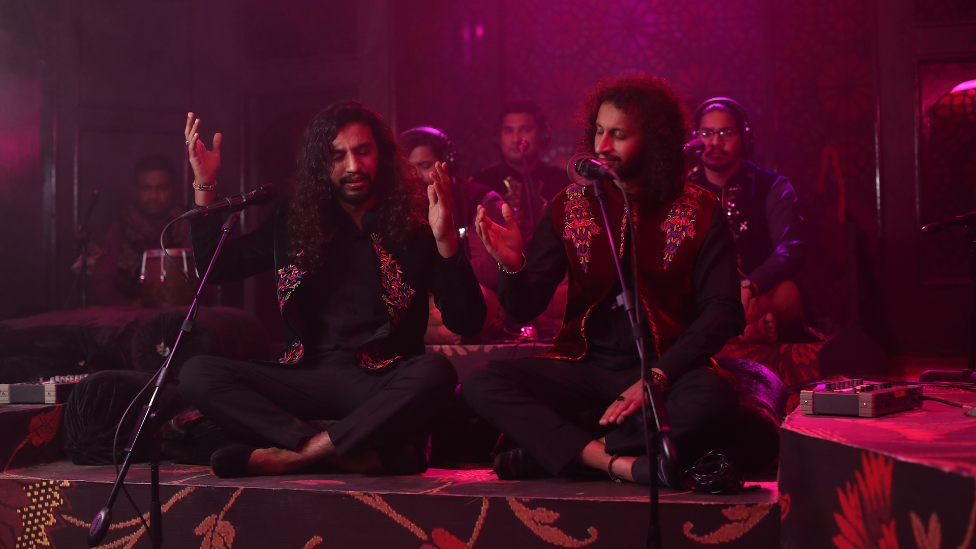 In a room lit by red and pink lighting, Zain Zohaib perform crosslegged on the floor with a group of musicians.