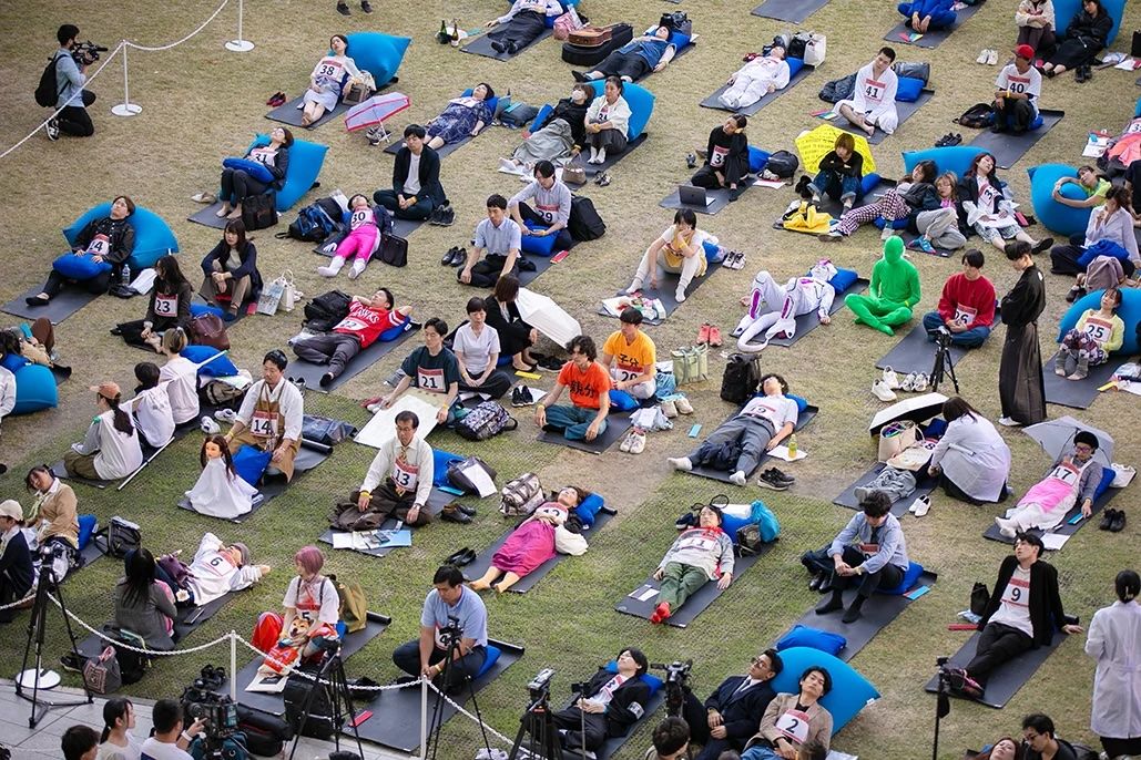 Bird's eye view of competitors lying on their backs on mats in a grassy field, staring into blank space