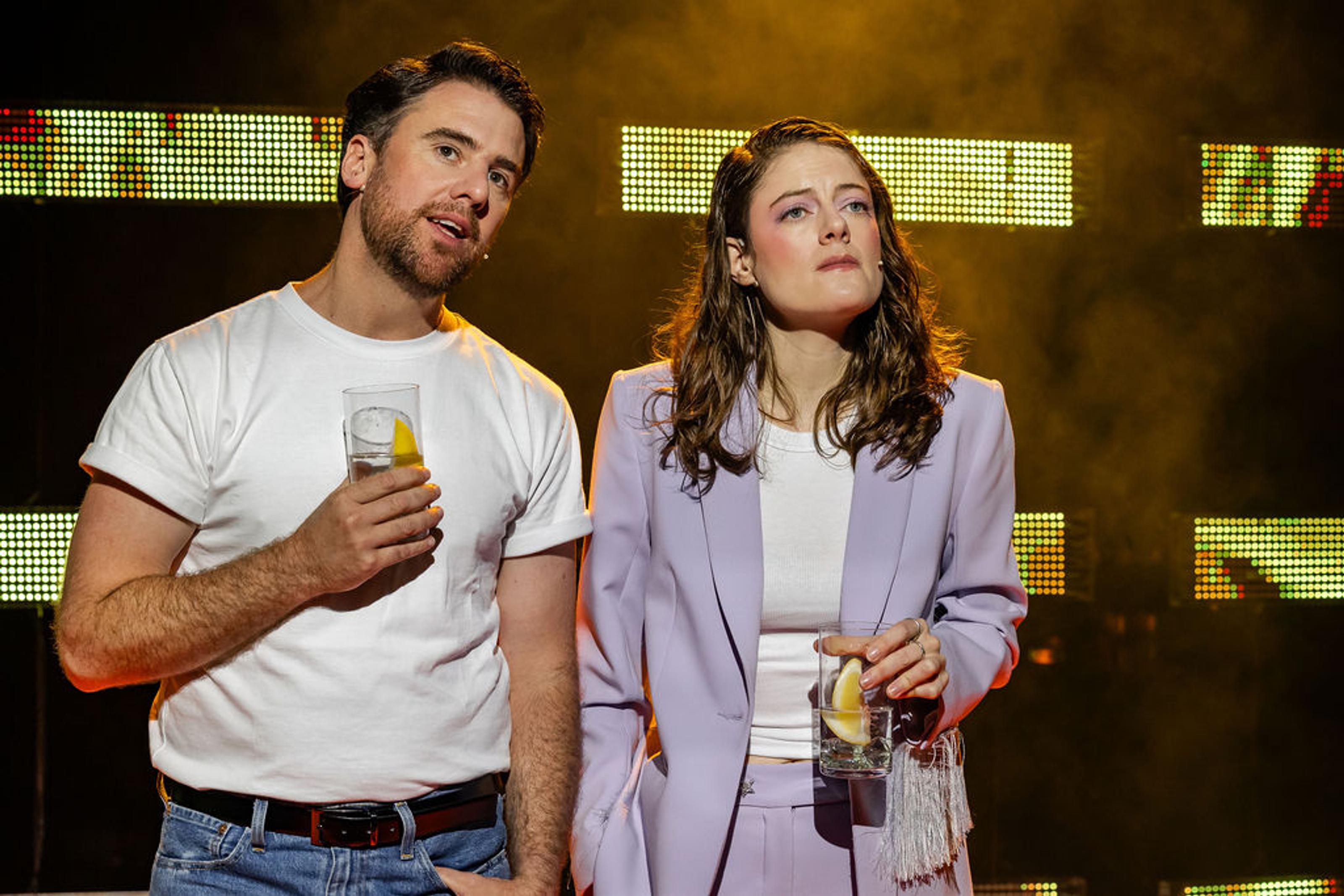 Two actors, each holding a drink, stand side-by-side looking forward at something out of frame