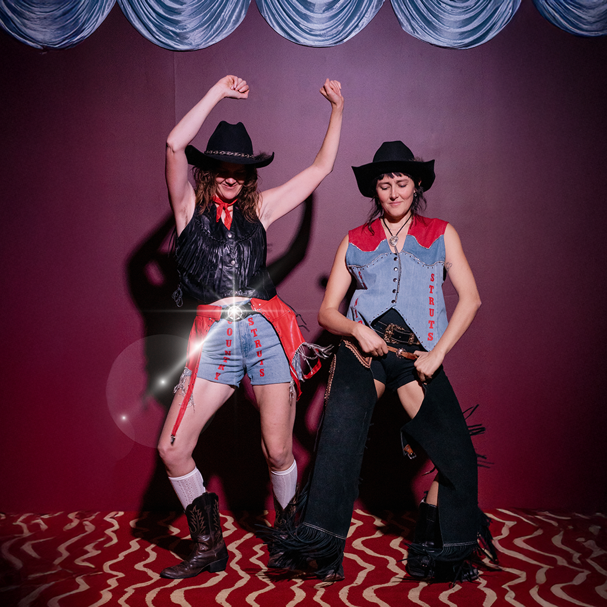 Dressed up in country attire, two dancers show off their moves—one with their arms in the air, the other gripping their belt.