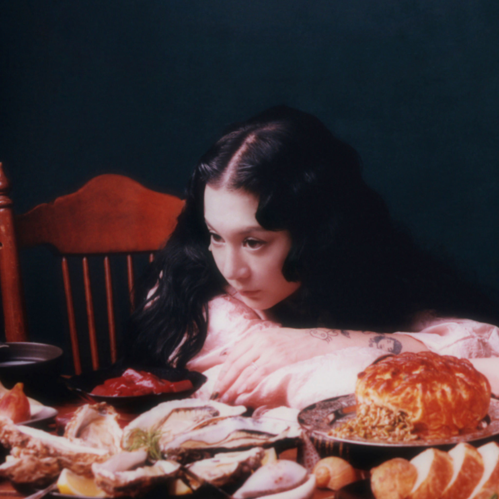 Japanese Breakfast lying down at a dinner table with a spread of food, flowers and a skull in front of her.