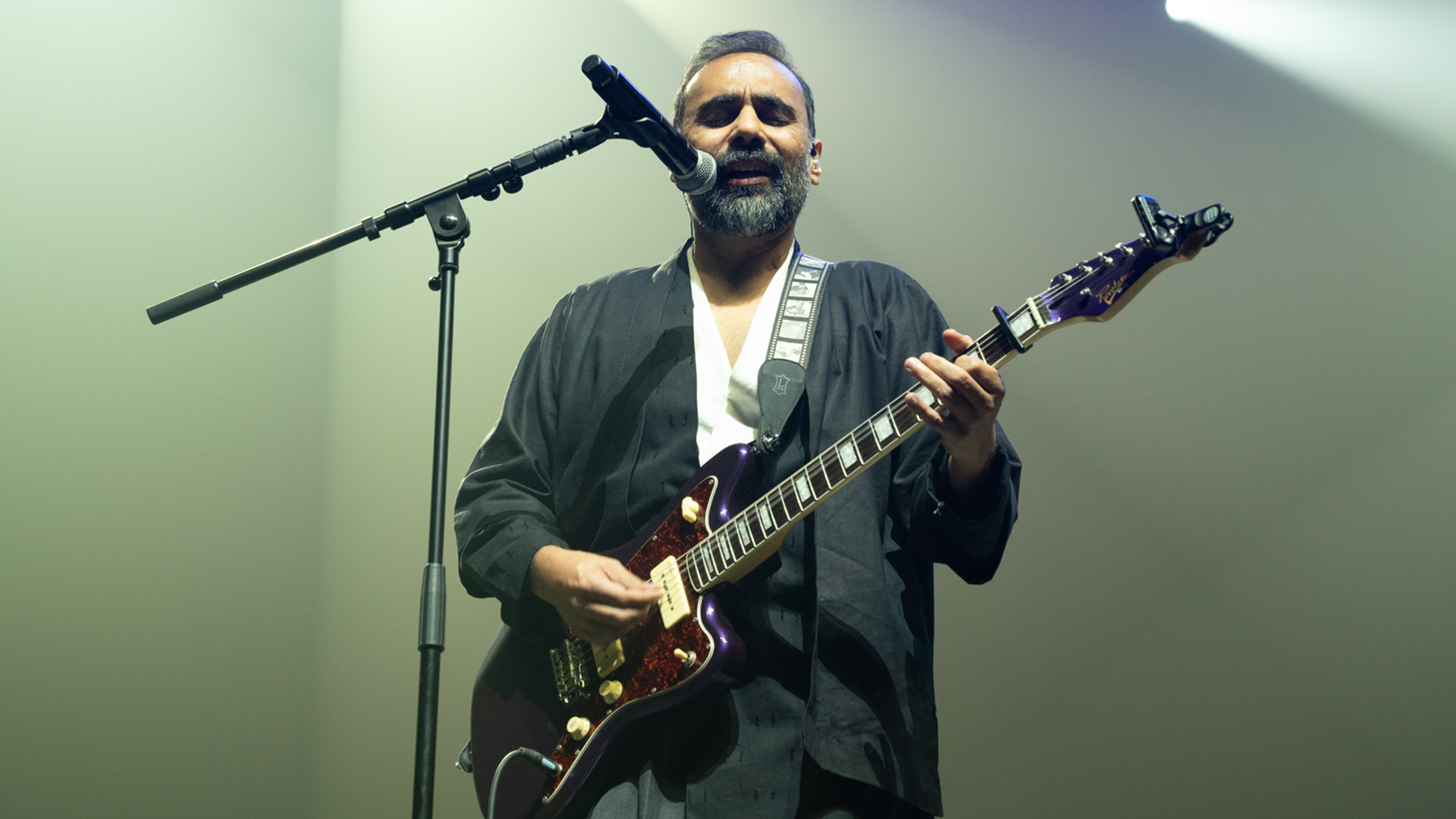 Lit by pale green lights, Xulfi sings into a microphone while playing the guitar.