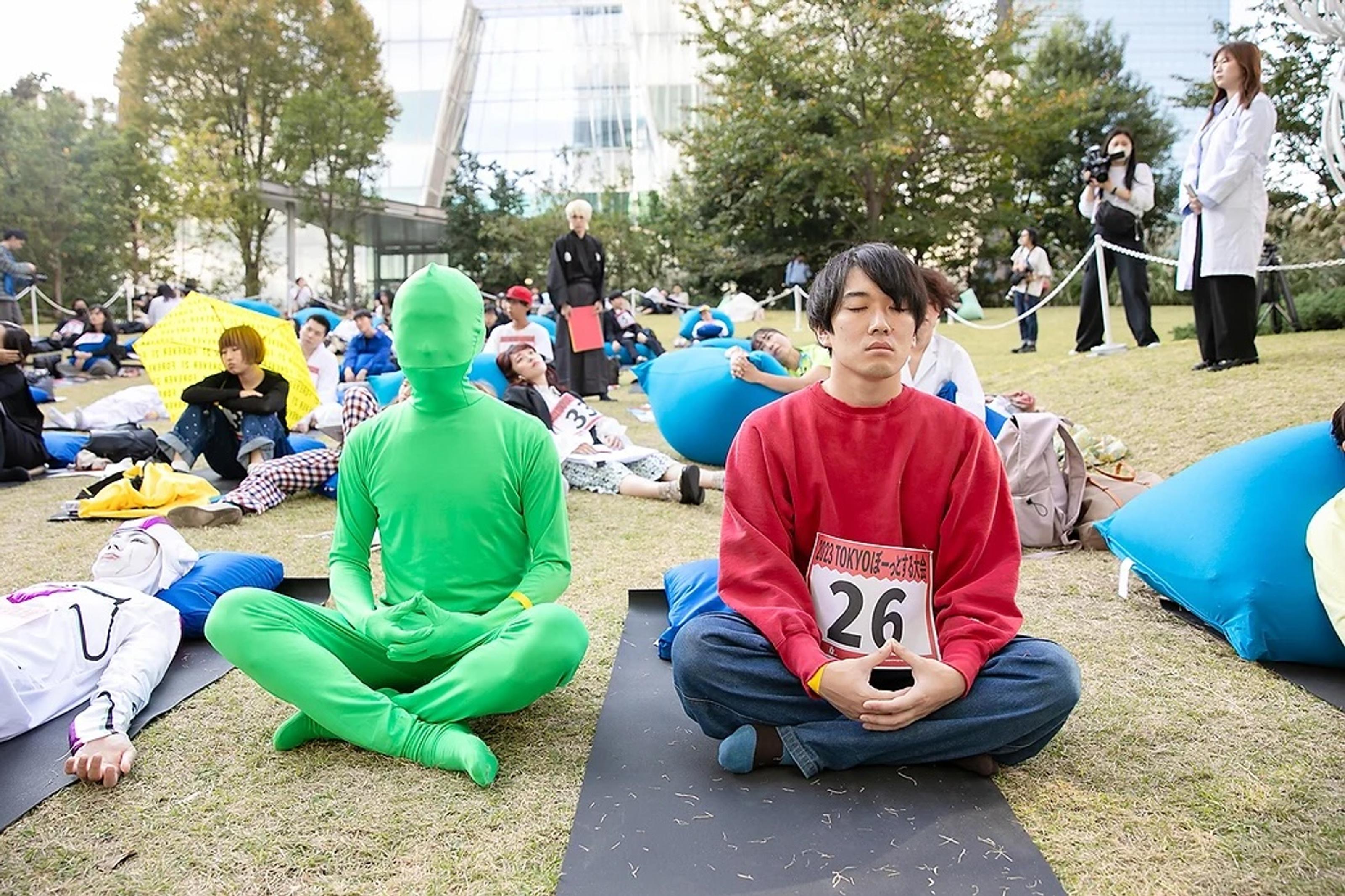 Two competitors, one in green body suit and the other in red sweater, sitting on the grass with their legs crossed