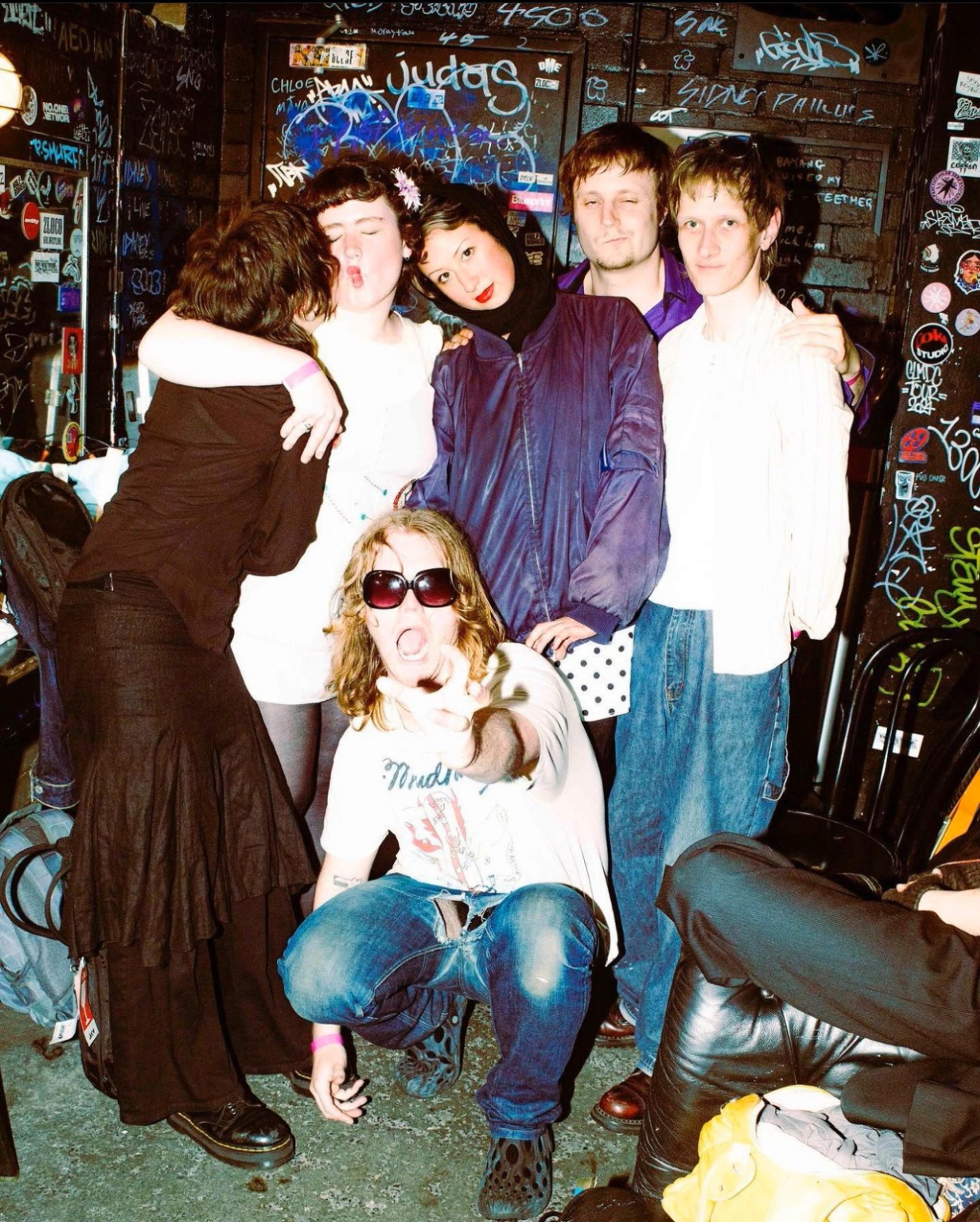 Six members of Spike Fuck posing together: one with sunglasses sits and points forward, the person on the far right hides their face in the next person's arm, the one in the center wearing a purple shirt rests her head on the second person from the left’s shoulder, and the second person from the right crosses his arm with the first person on the right.