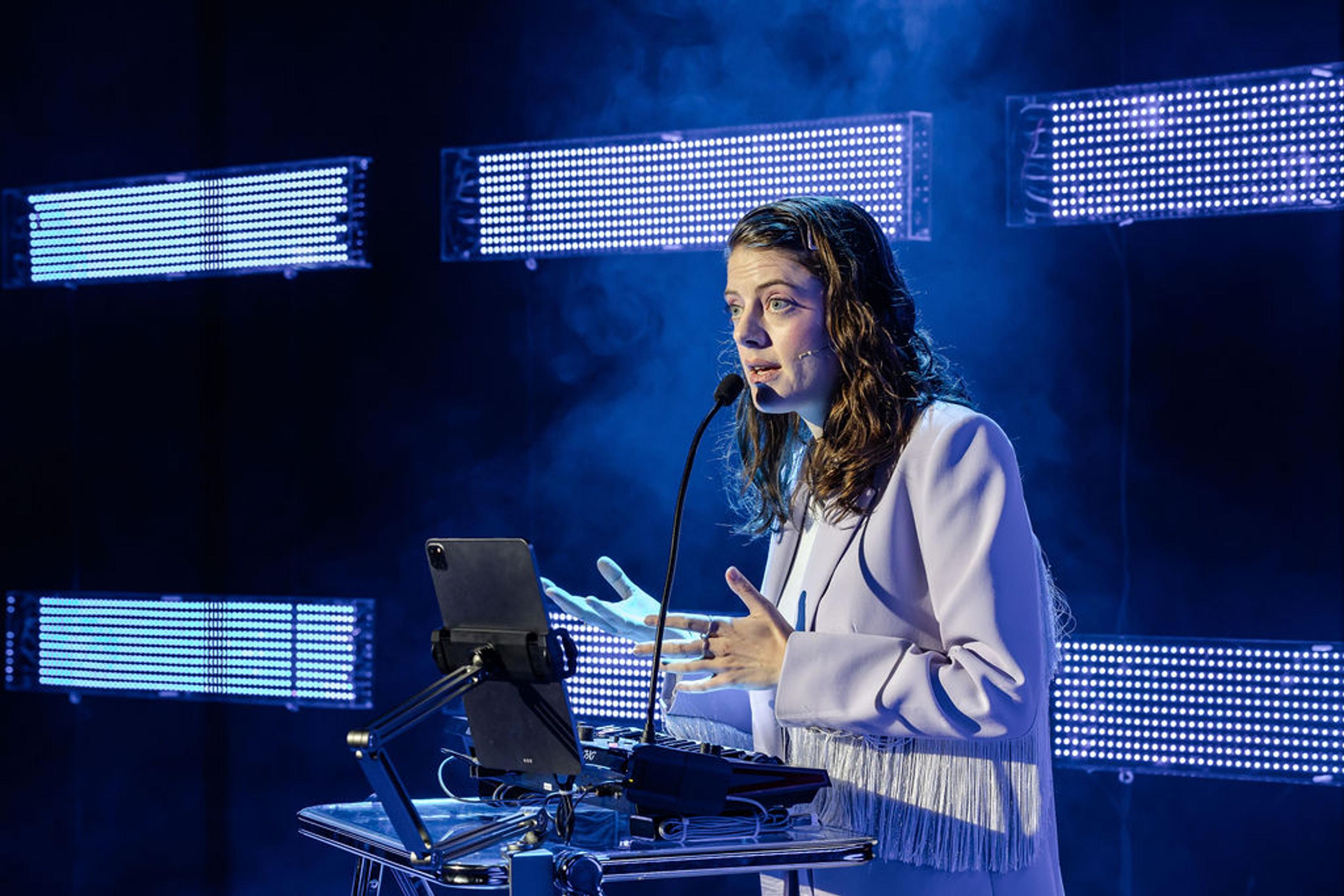 Karin McCracken mid-performance lecture, standing at a lectern with a screen
