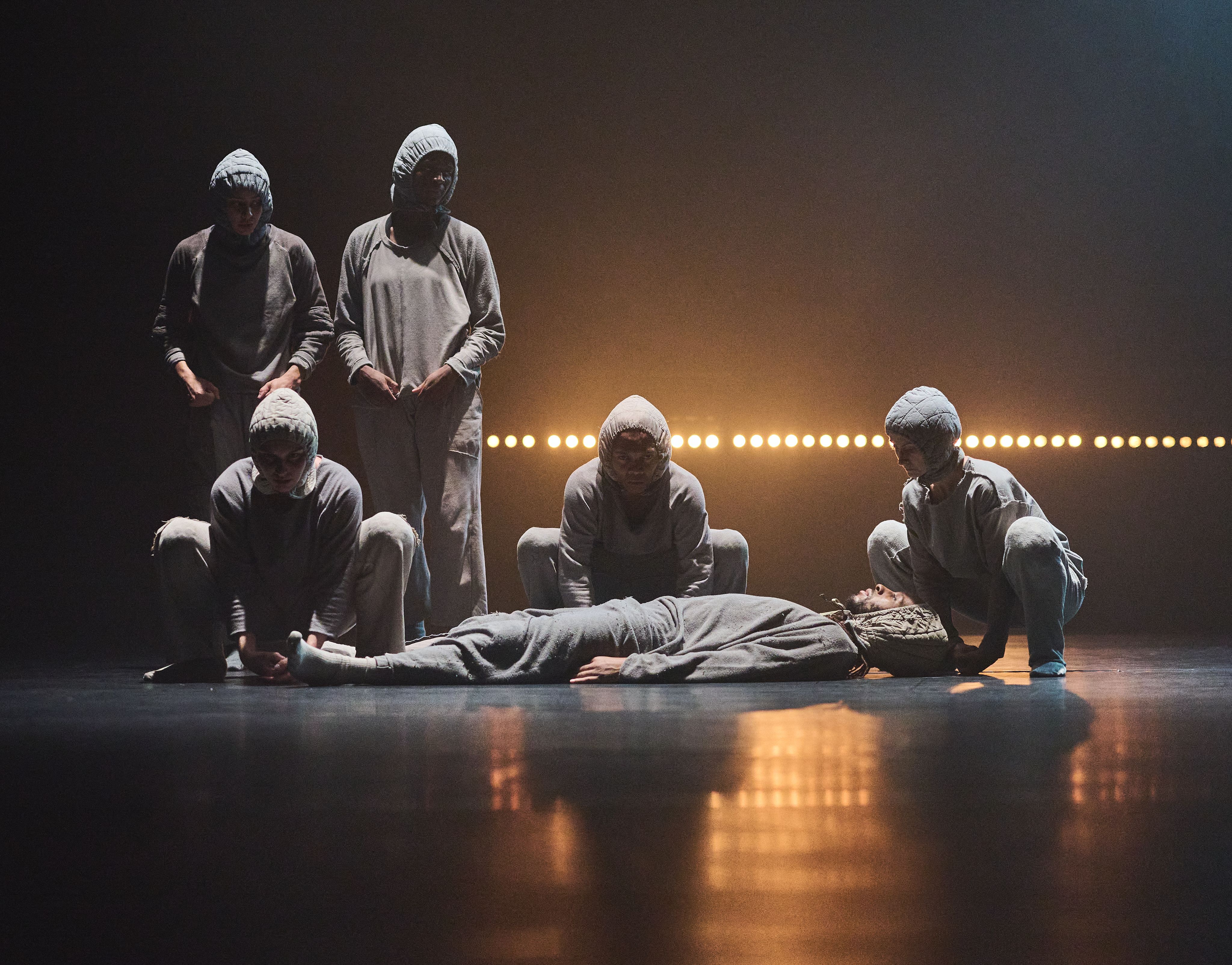 Five dancers in hoodies, one lying on the floor with eyes closed and the other four surrounding them.
