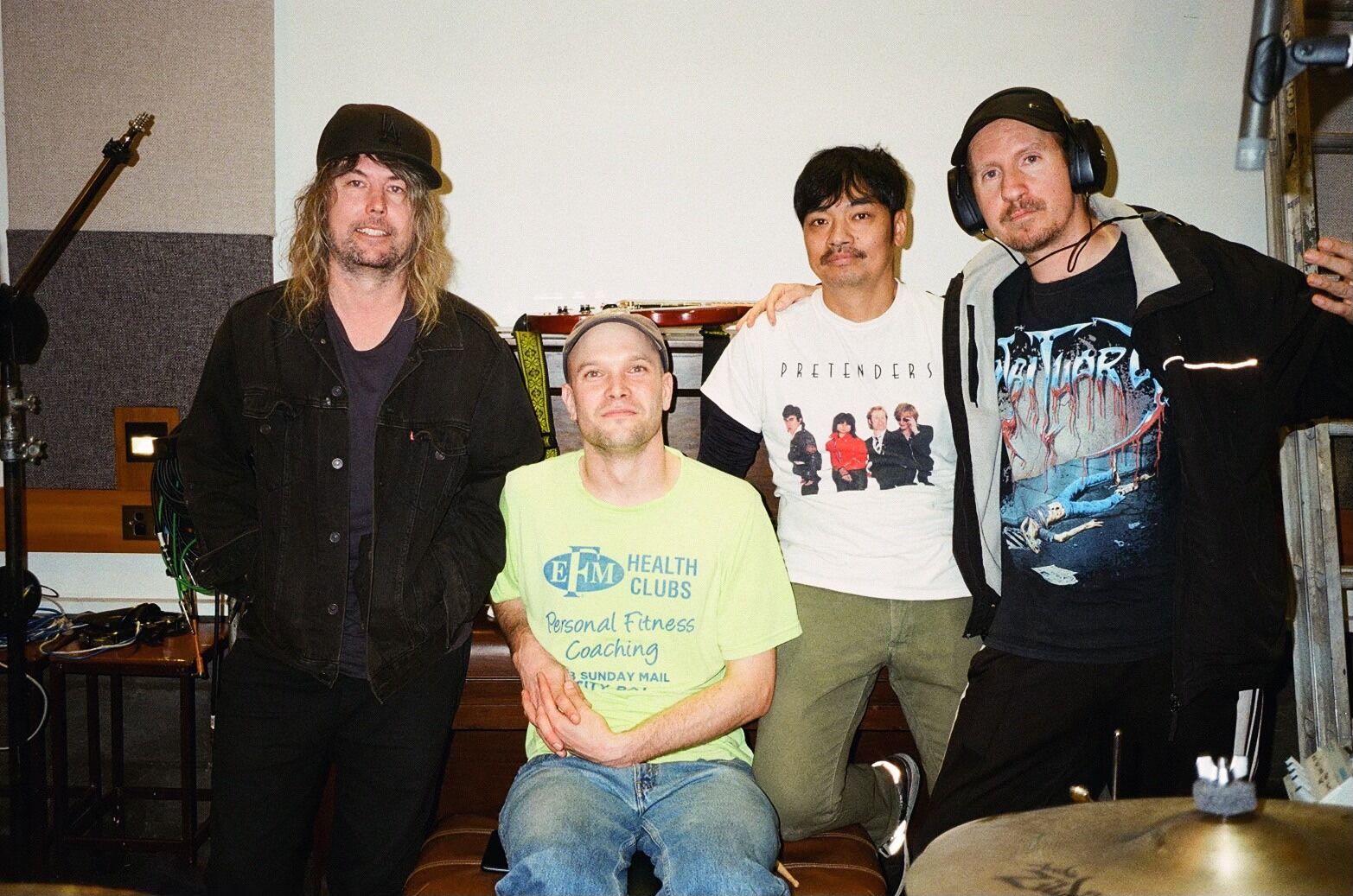 In the group of four, three people are standing. From right to left: the first person is wrapping their arm around the second person's shoulder. The third person is seated, while the fourth person is standing and smiling. 