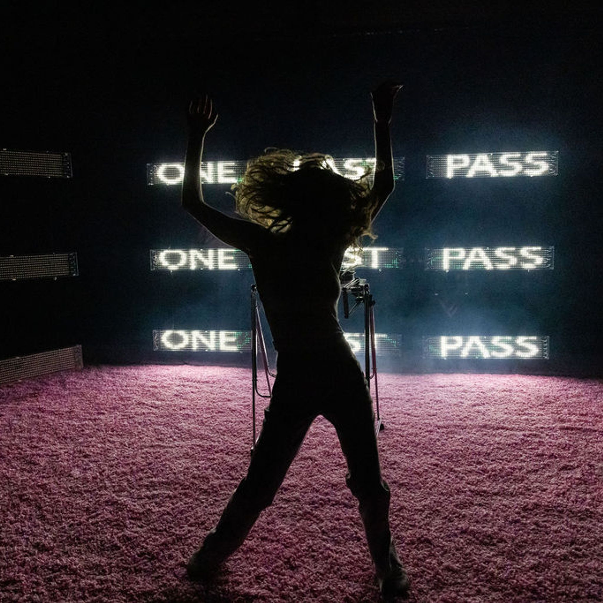 A silhouetted body mid-dance in front of LED screens that read “One last pass”