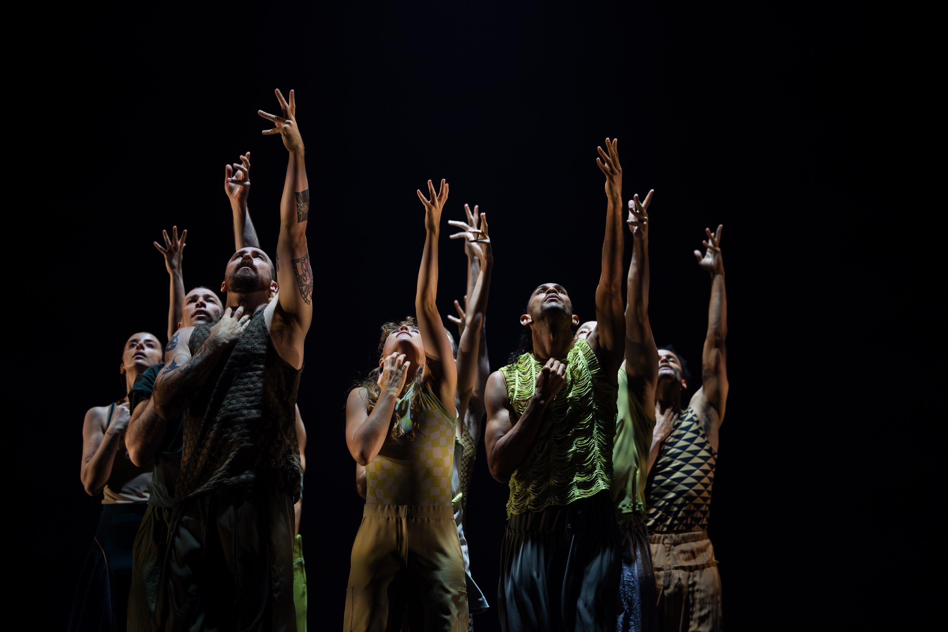 Several dancers stand up, looking to the roof with their left arm stretched upwards and their right hand in front of their chest.