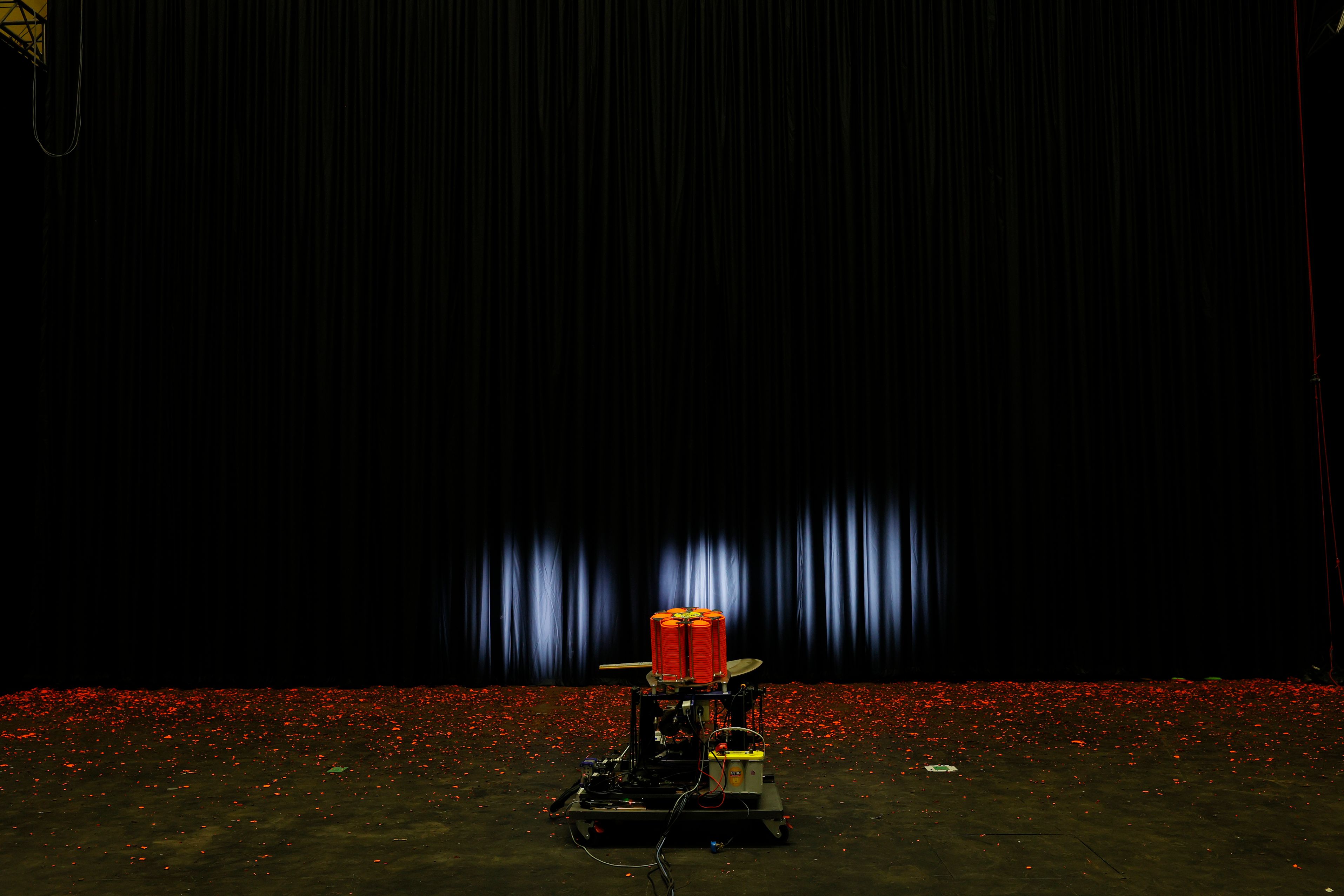 A trap machine is placed on black wooden floor in front of a curtain. In front of the curtain are orange fluroescent pieces of broken clay targets. Three spotlights in circular shapes shine on the large black curtain. 
