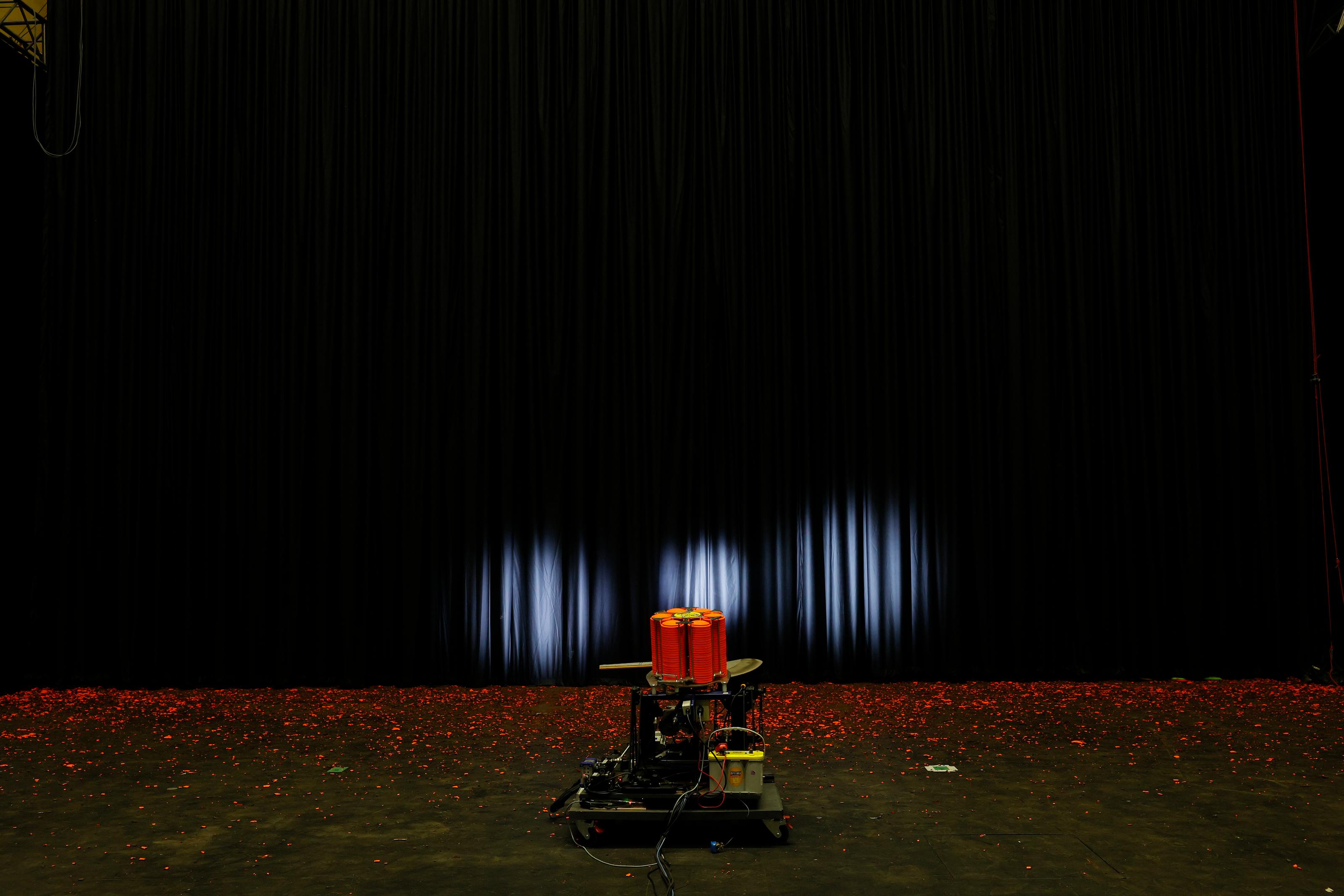 A trap machine is placed on black wooden floor in front of a curtain. In front of the curtain are orange fluroescent pieces of broken clay targets. Three spotlights in circular shapes shine on the large black curtain.