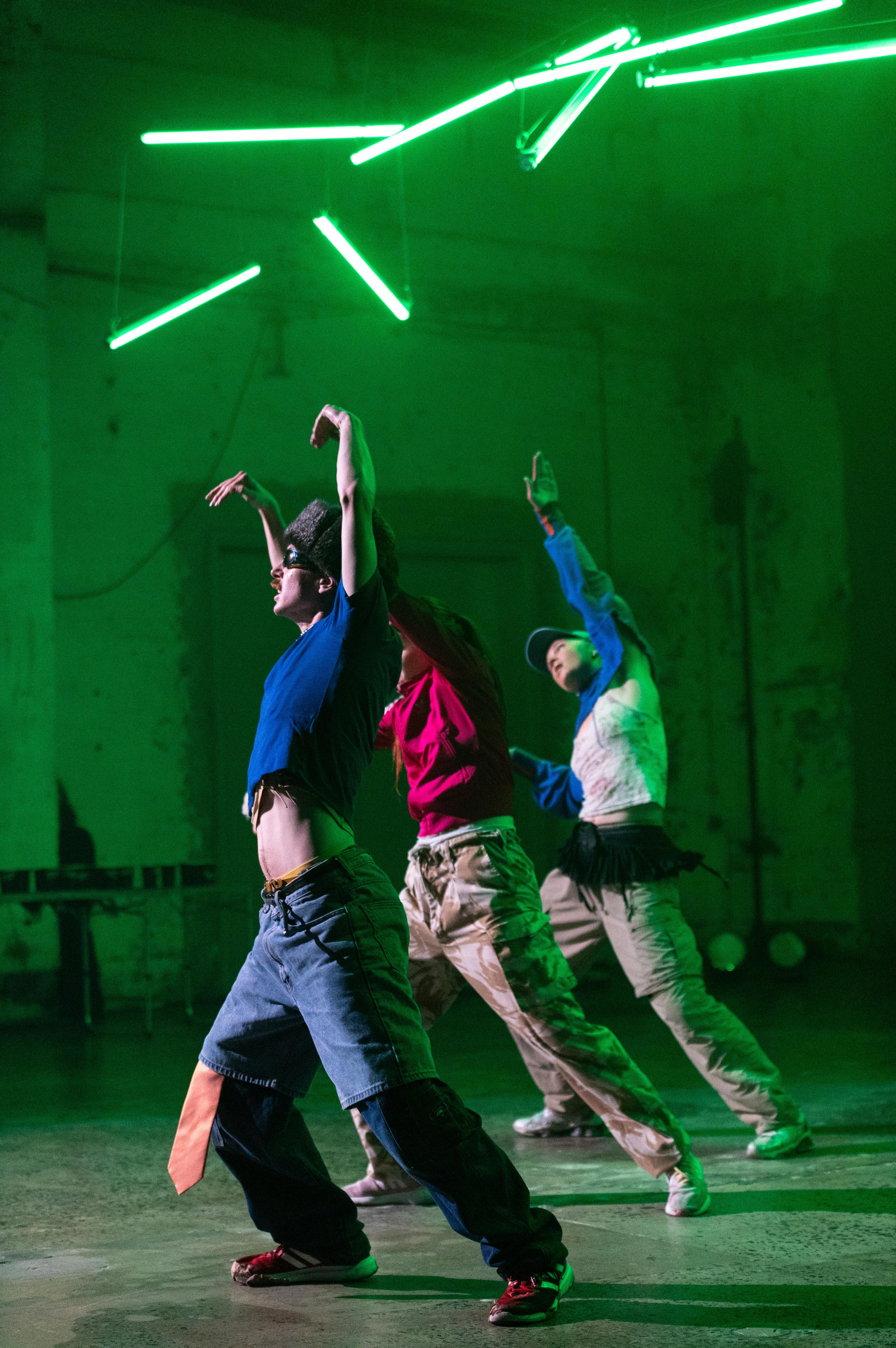 A trio of dancers pose with their arms in the air and with one of their legs extended out, under hanging light rods.