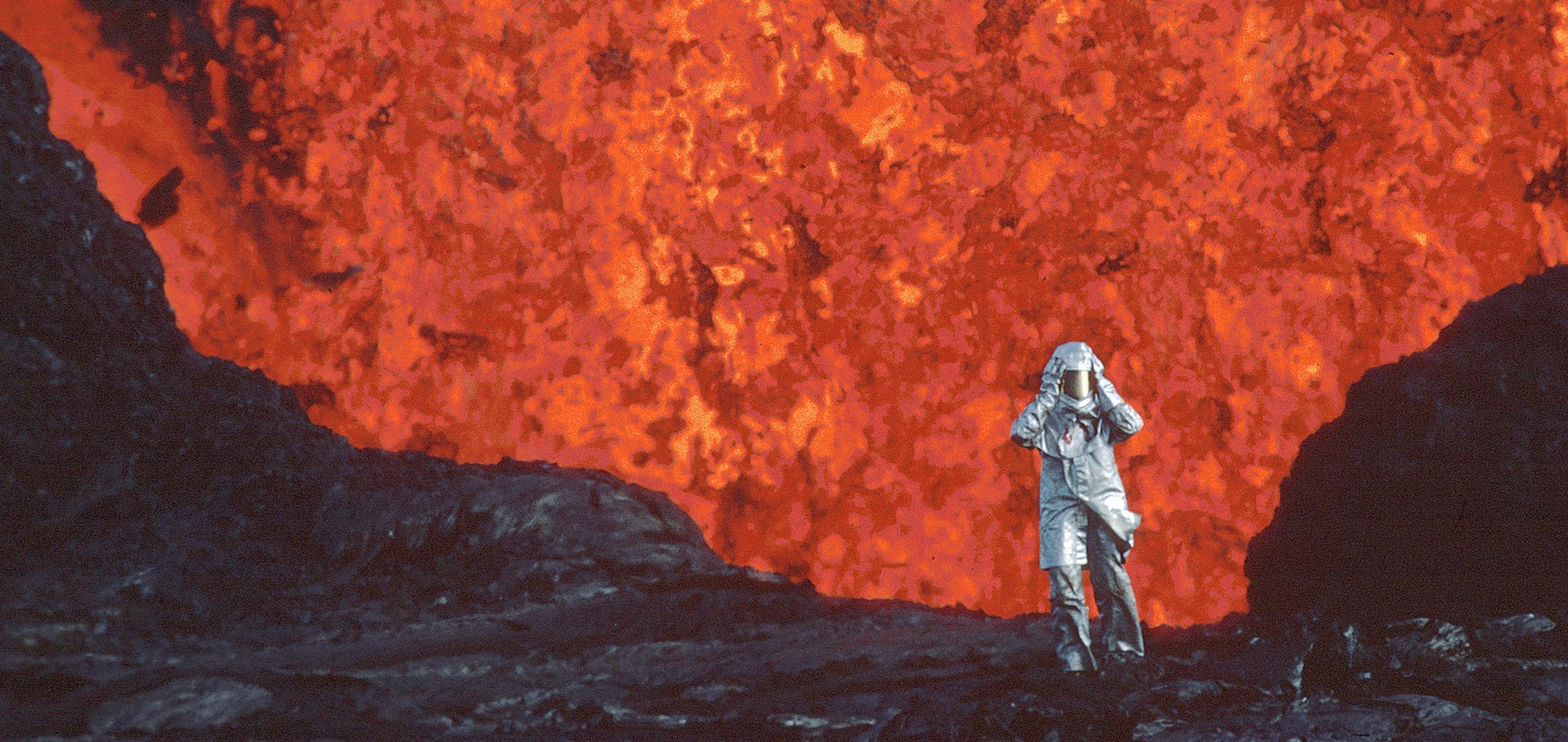 A person in a heat-protective suit stands on dark volcanic rock, facing a massive wall of flowing lava.