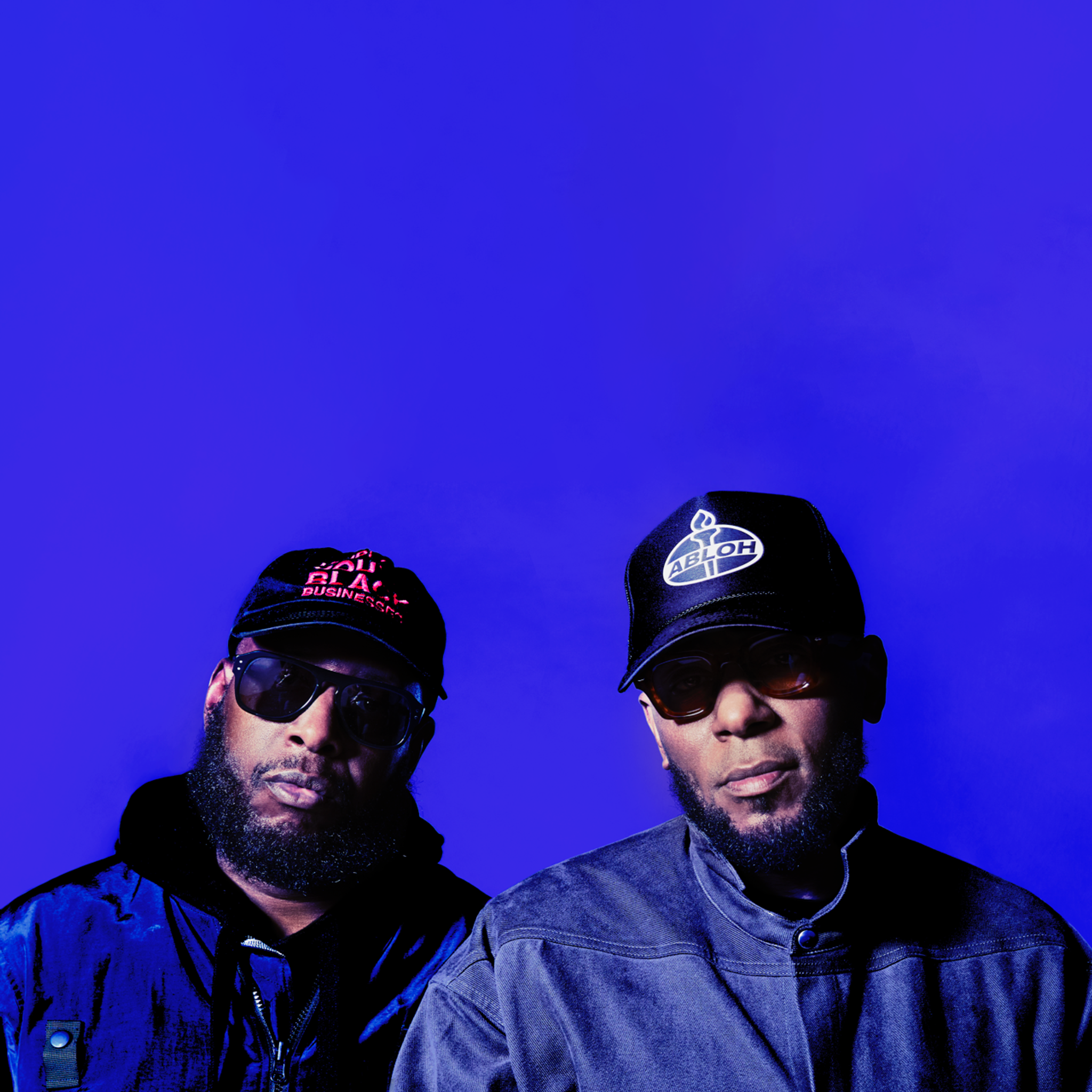 Two bearded hip hop stars are standing next to each other. Both wearing caps and sunglasses.