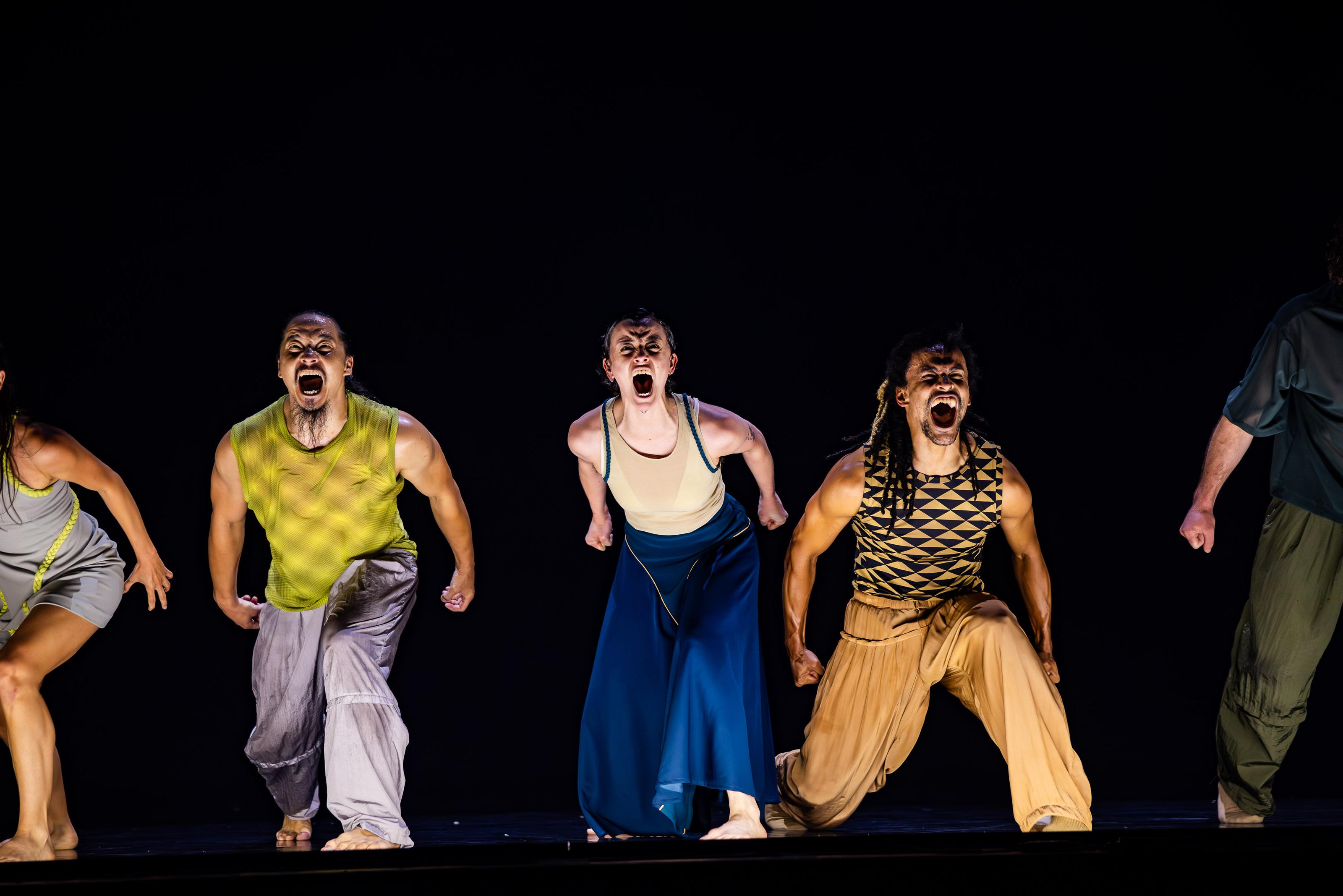 Three people lunge towards the audience and camera with their arms behind them, shouting.