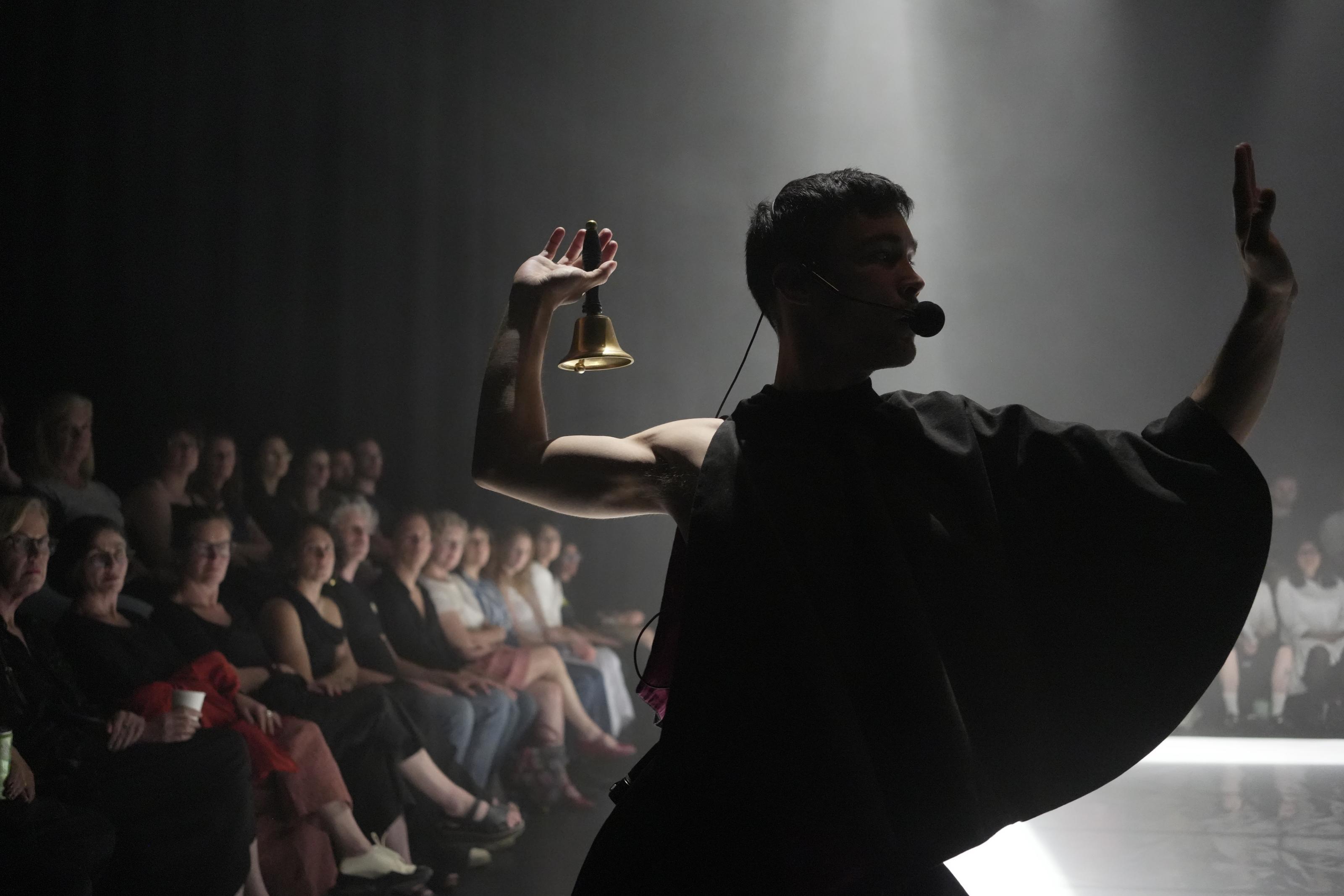 Silhouette of a performer wearing a mic and draping outfit, posing in the foreground with a bell, as the spotlight behind them illuminates the seated audience in the background.