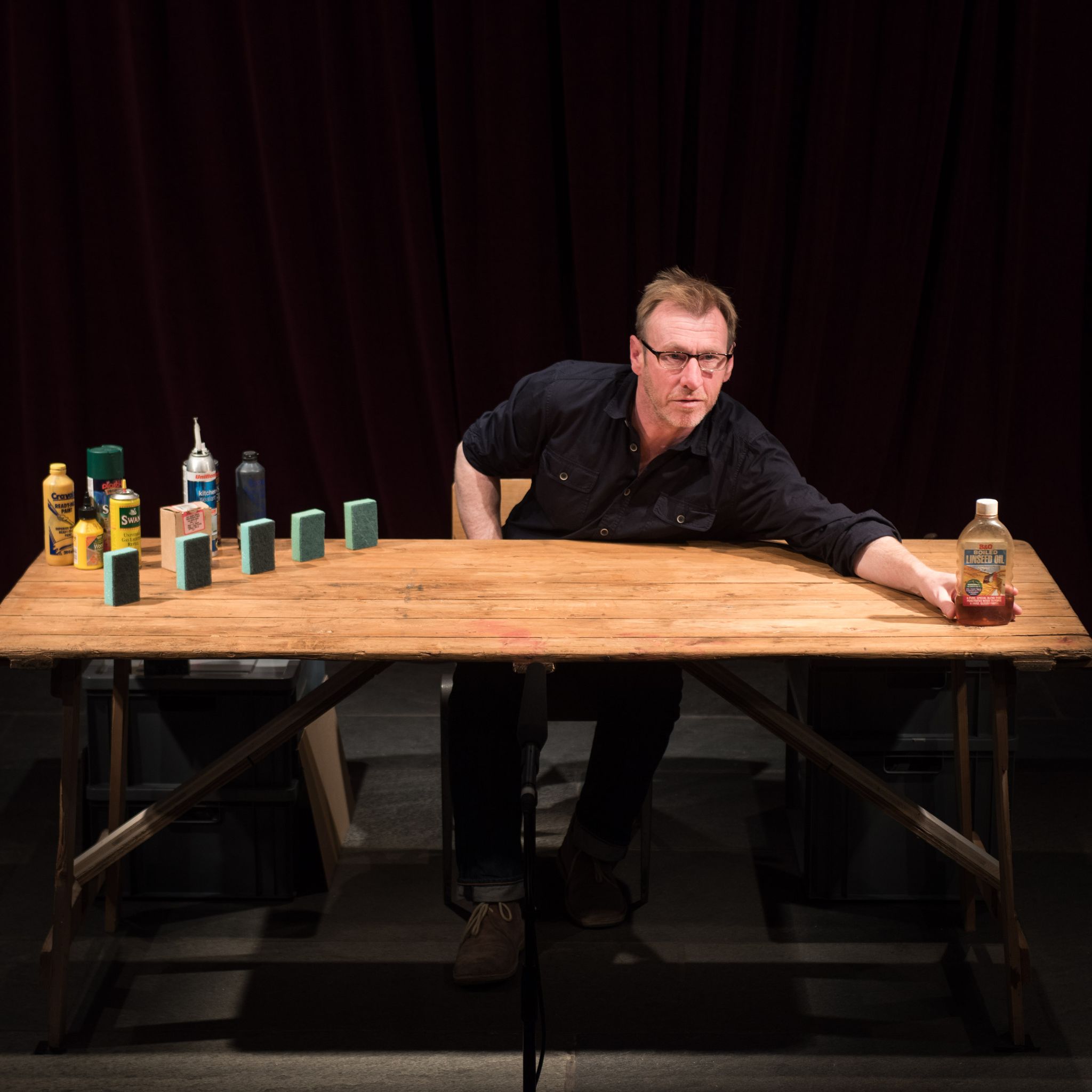 An actor sits at a wooden table with bottles, jars and sponges lined up on the left side. One singular bottle is being pushed to the right by the actor.