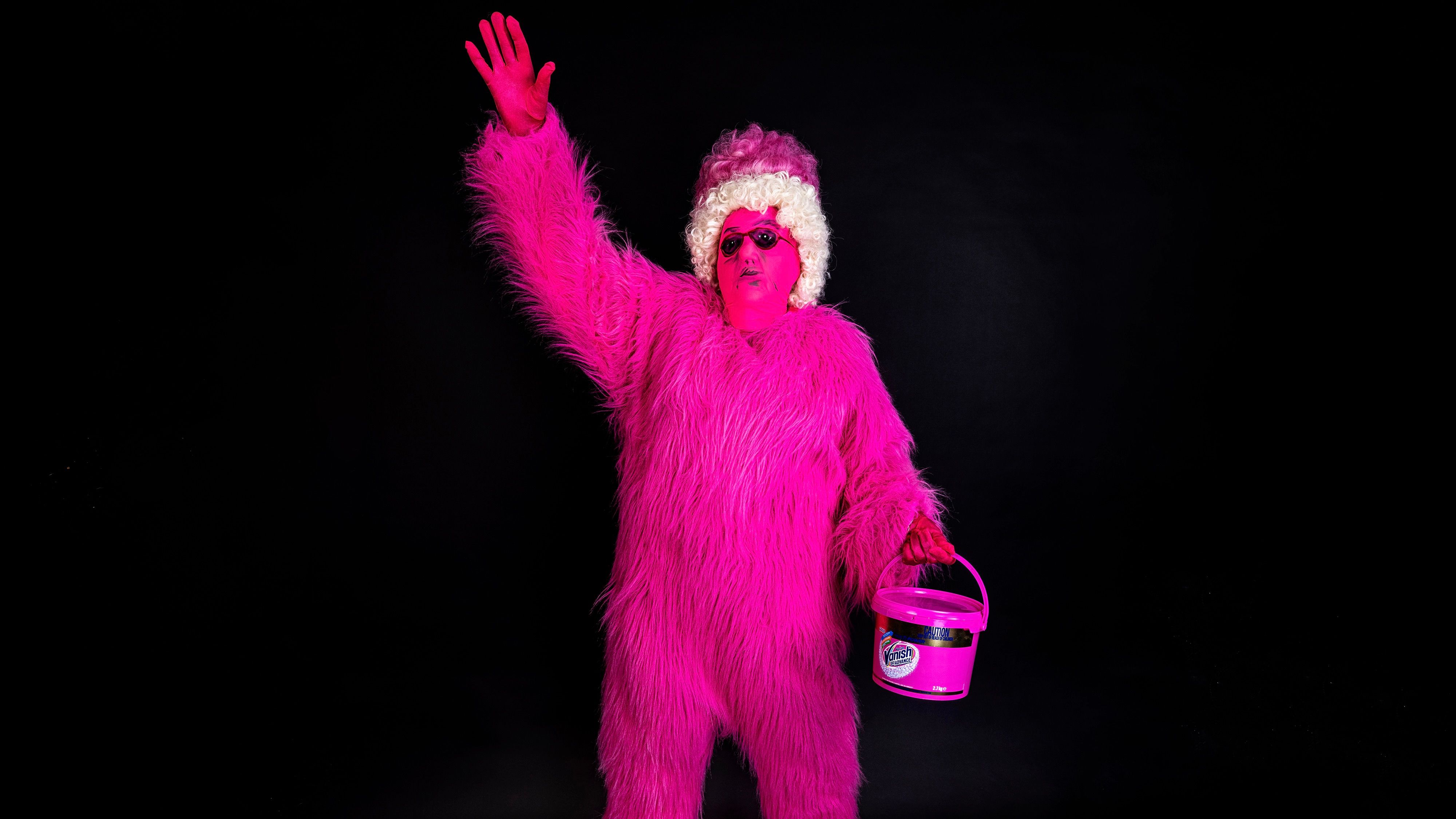 Person dressed in a vibrant pink furry consume with one arm held in the air and the other holding onto a Vanish bleach bucket. Face is covered by a pink mask with black marker sunglasses, on their head is a curly pink and white wig. 