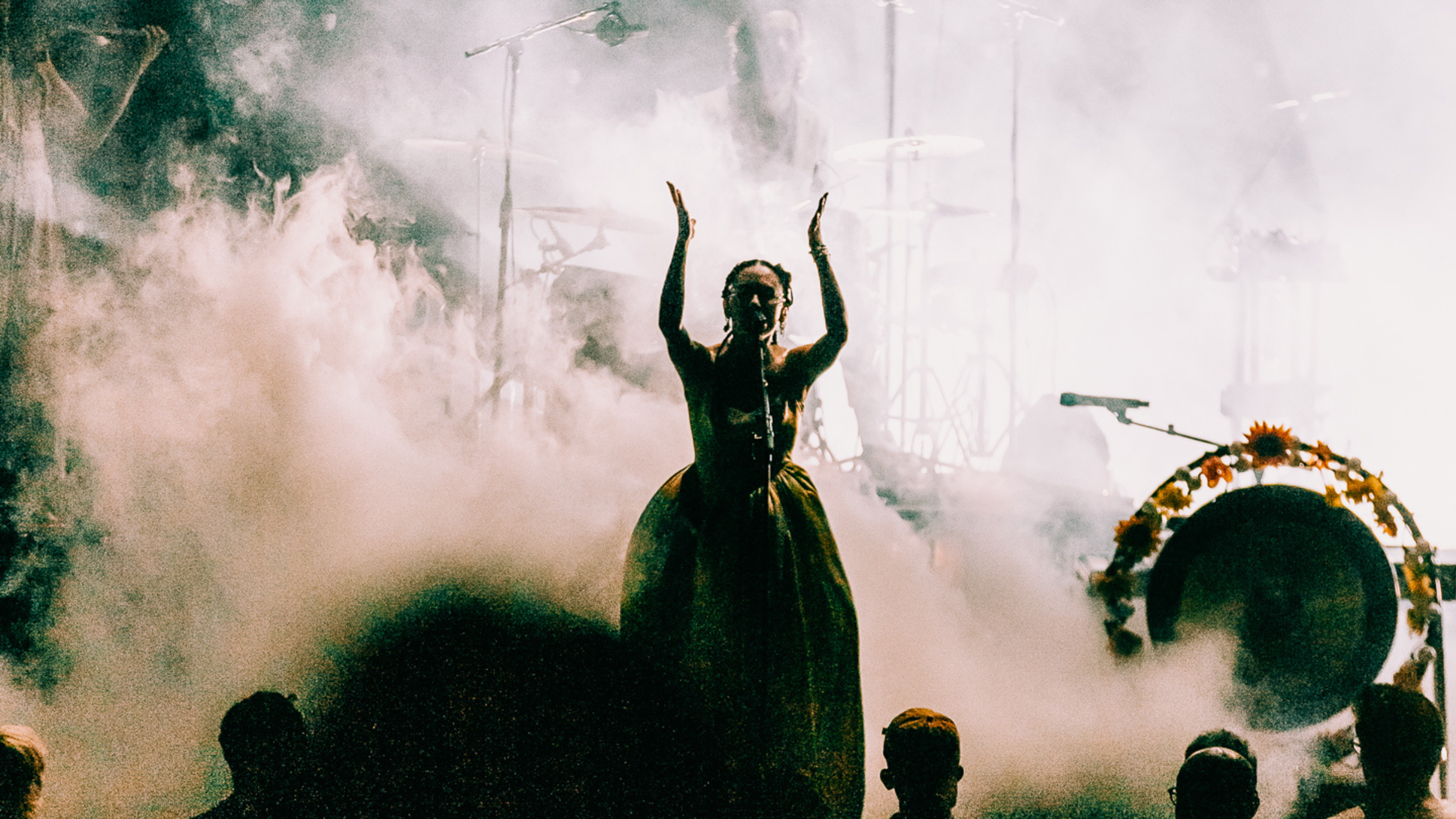 Japanese Breakfast performs on stage, singing into a microphone with raised arms. The lighting almost creates a silhouette of the performer against a hazy background.