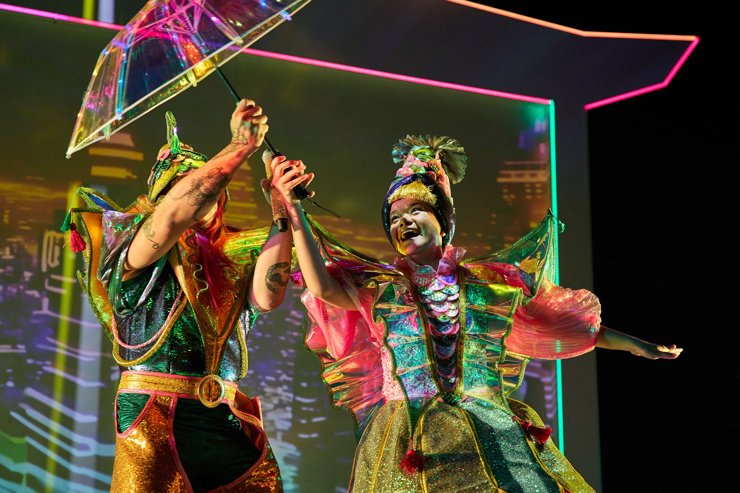 Two people in over-the-top neon Ancient Chinese style clothing hold and umbrella