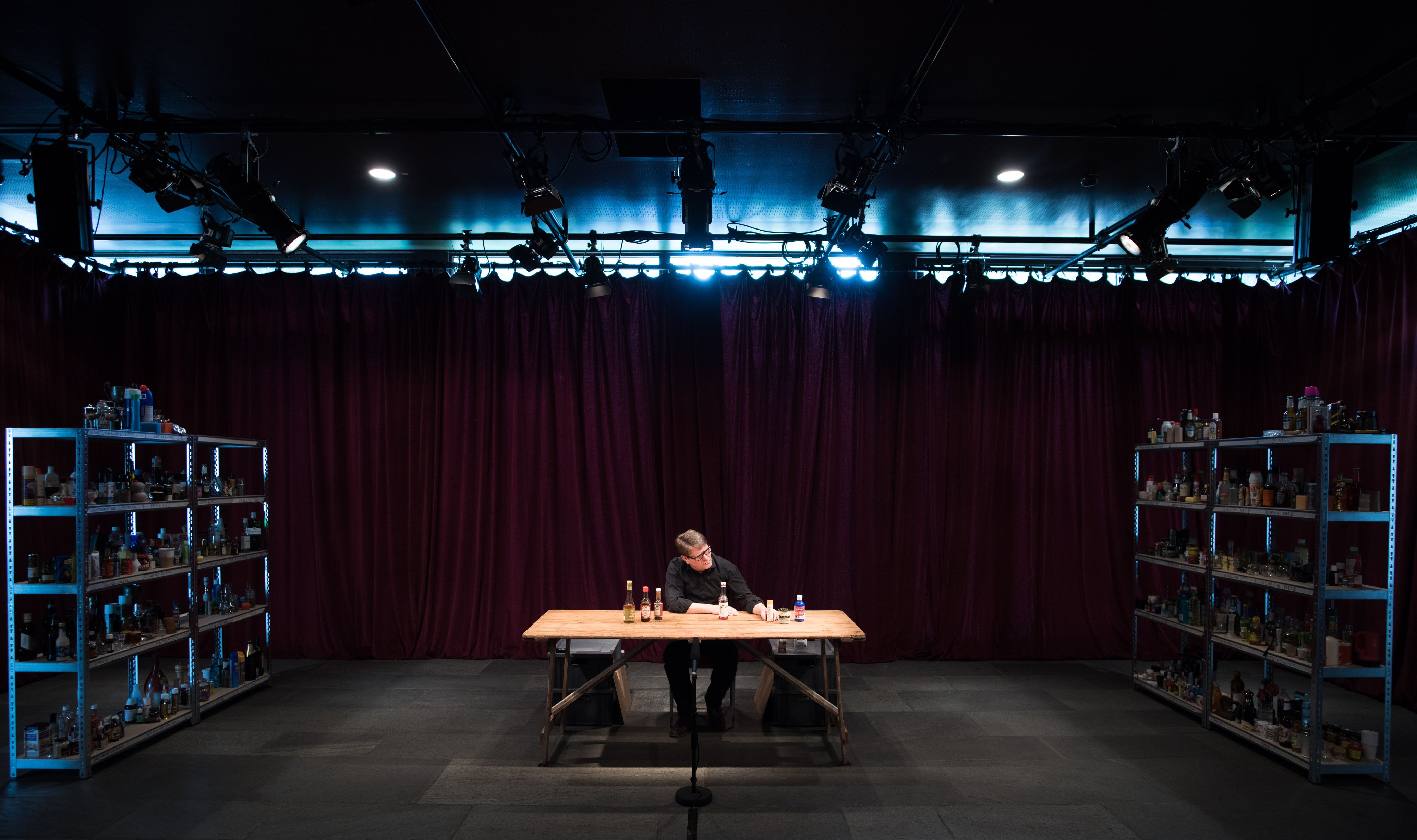  An actor sits in the centre of a room at a large wooden table holding various bottles and jars. Shelves of similar objects are positioned on either side of him.    