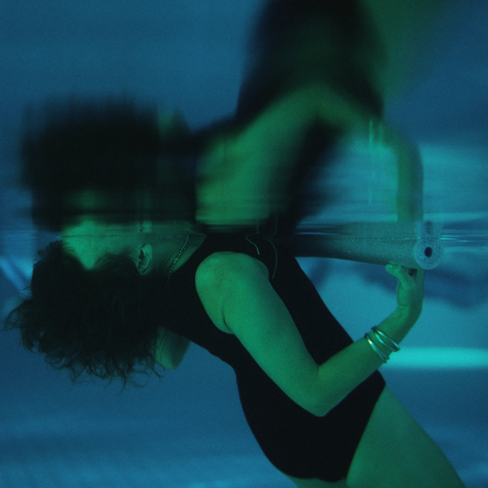 An underwater photo of a person floating backwards in a pool with their head submerged, holding onto a pool noodle.