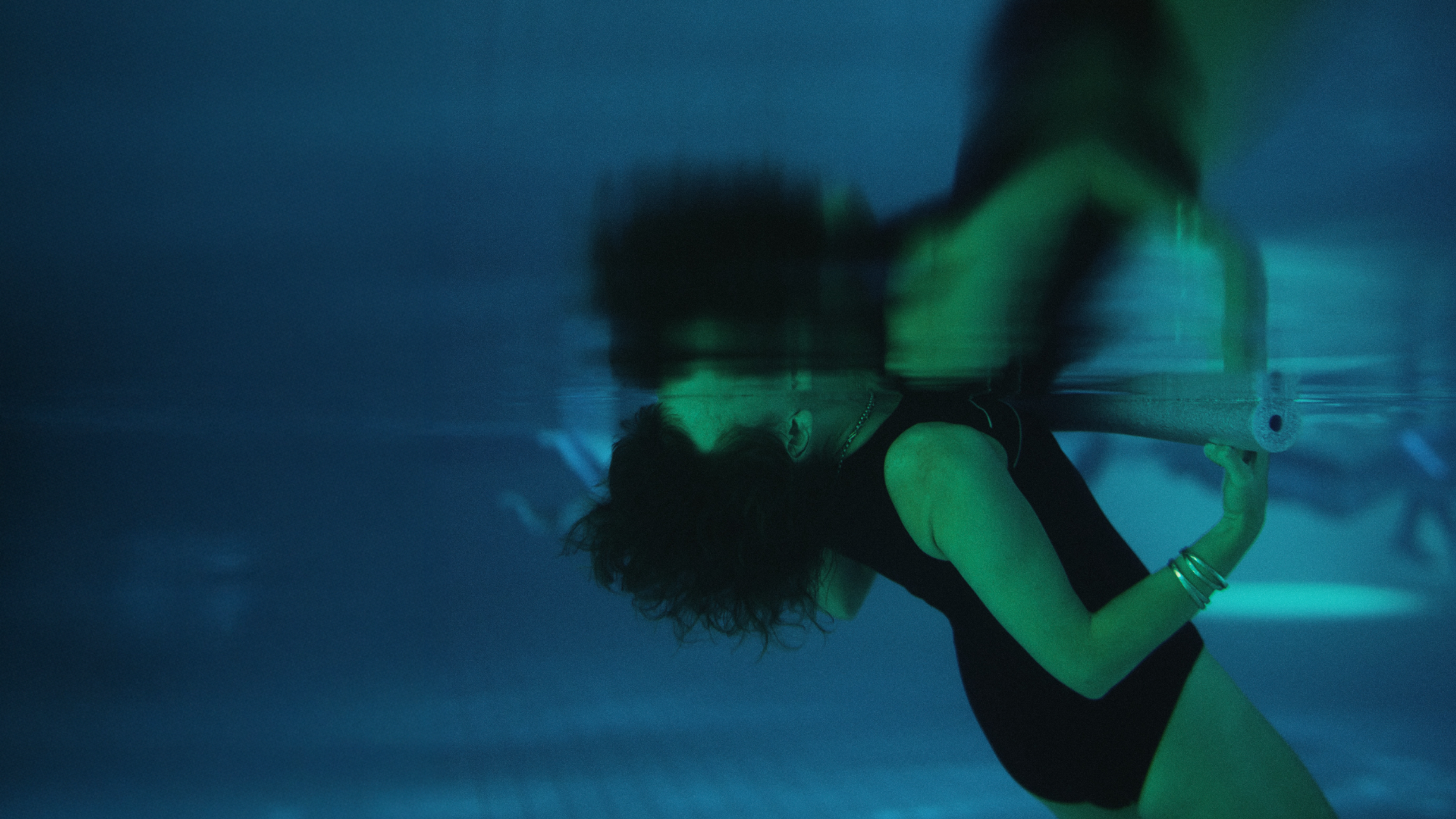 An underwater photo of a person floating backwards in a pool with their head submerged, holding onto a pool noodle.