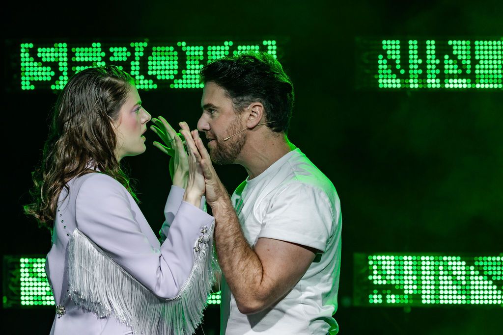 Two actors face each other, hands raised and interlocked, in front of an LED screen that reads “serotonin” 