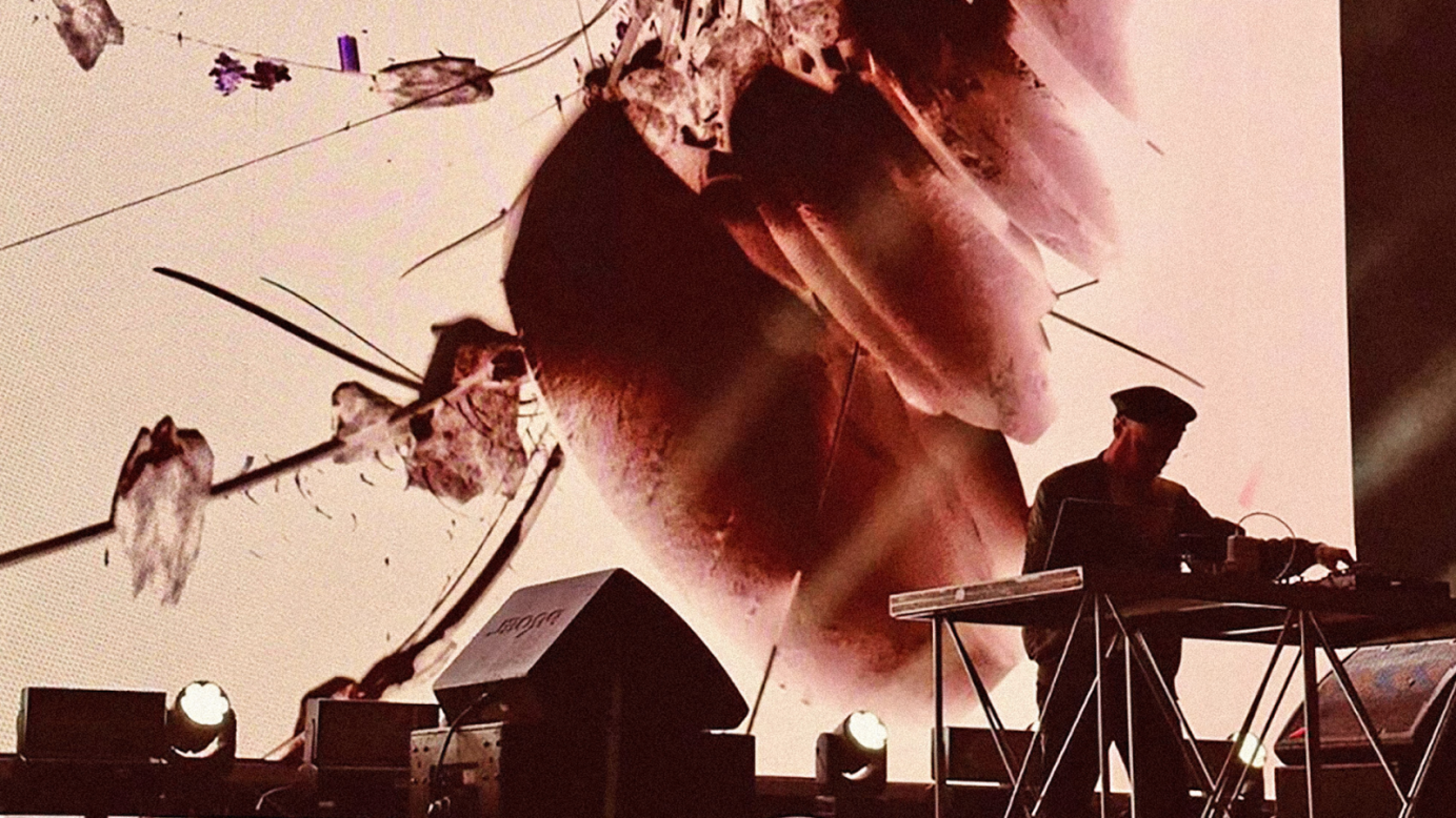 A performer operates a sound equipment in front of a large screen with abstract visuals, illuminated by stage light. 