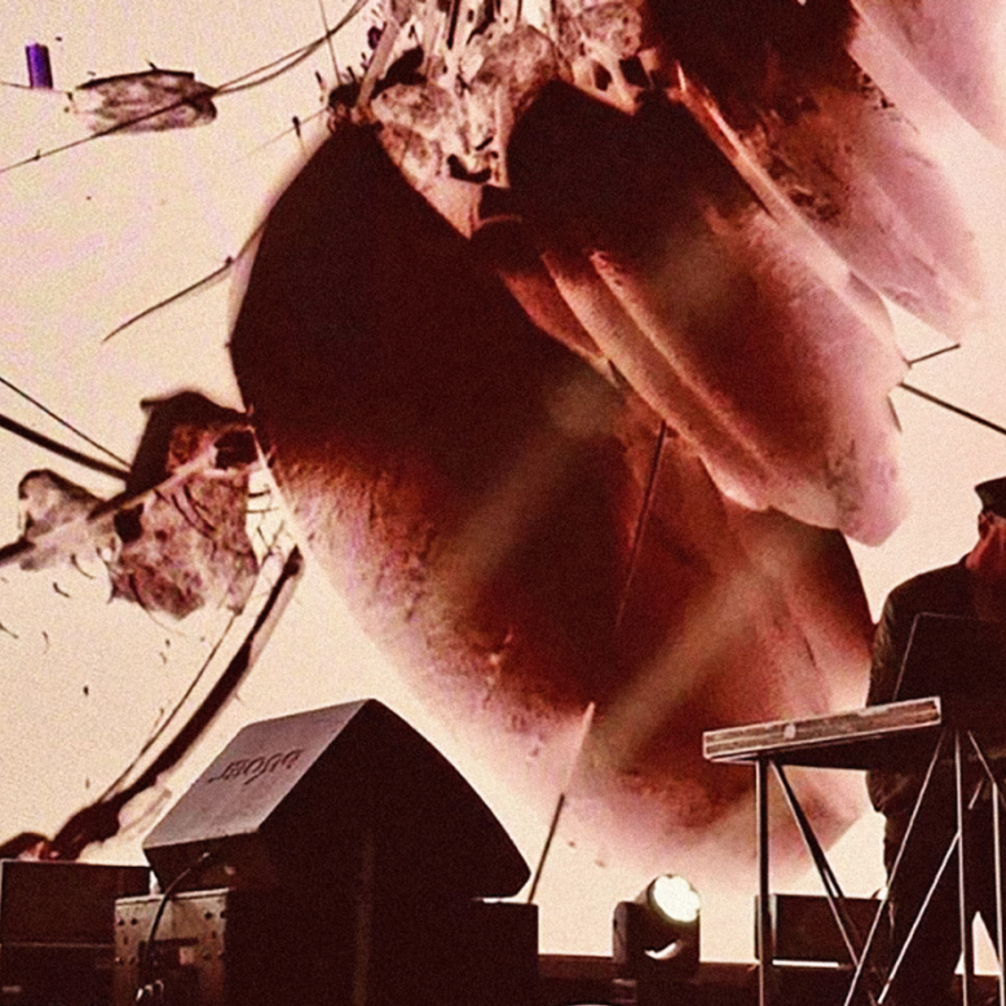A performer operates a sound equipment in front of a large screen with abstract visuals, illuminated by stage light.