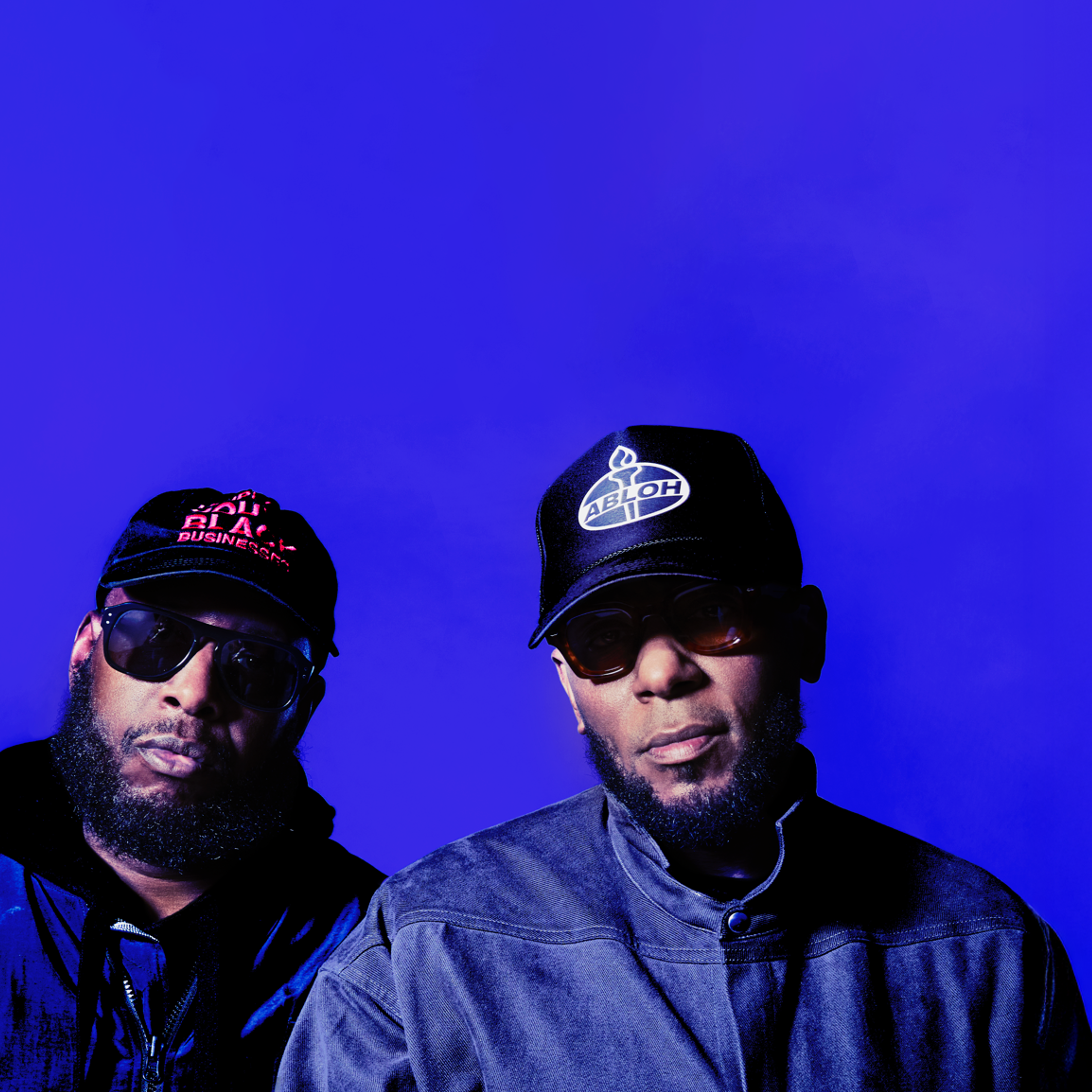 Two bearded hip hop stars are standing next to each other. Both wearing caps and sunglasses.