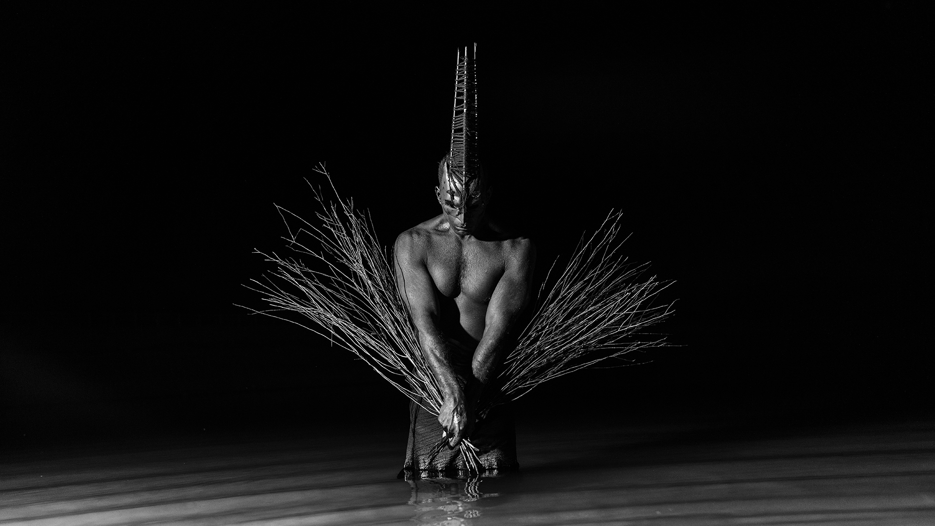 Kalaji kneels on the floor holding sheefs of wheet and wearing a mask