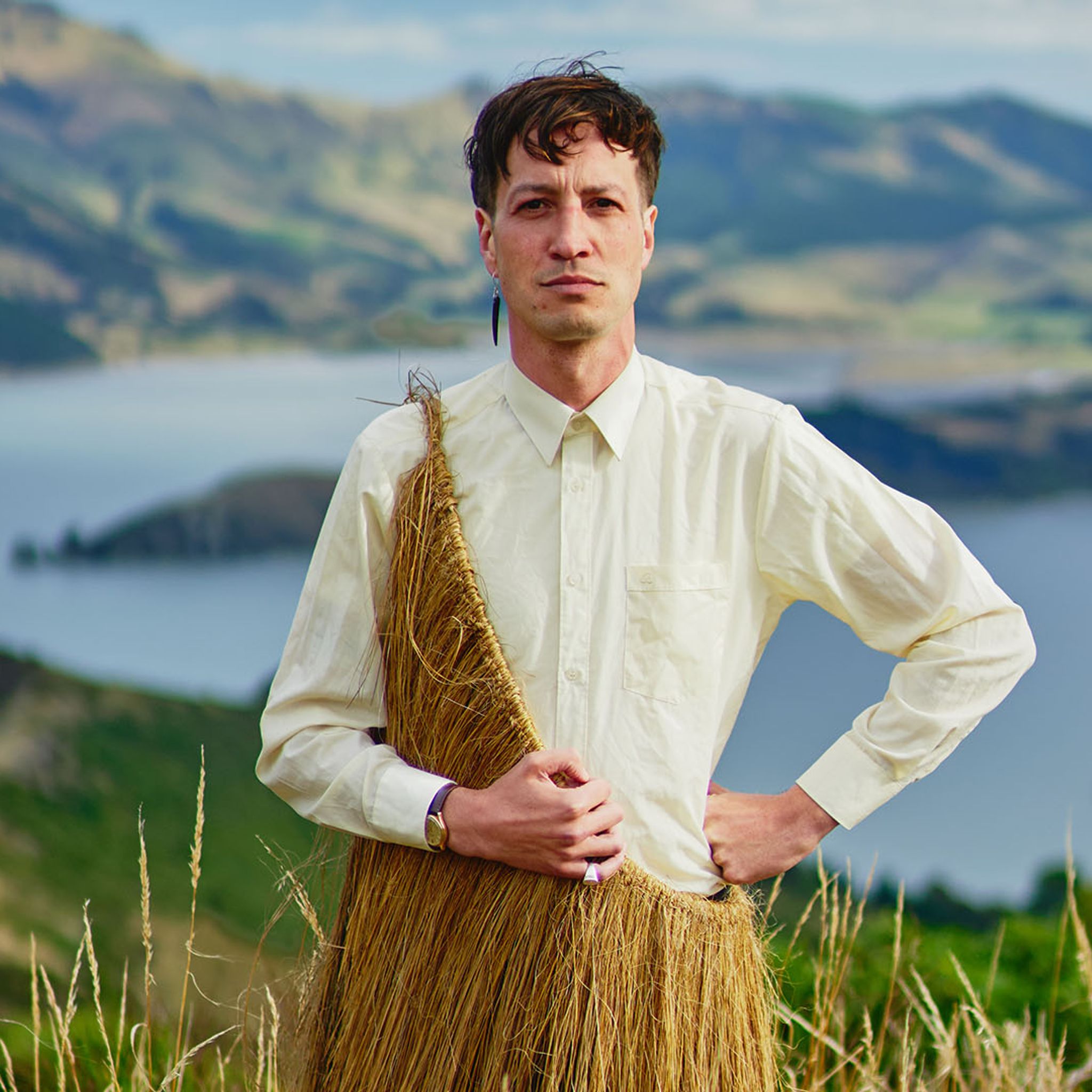 Marlon Williams stands in the foreground wearing a Pākē in front of a sweeping landscape of a bay and mountains