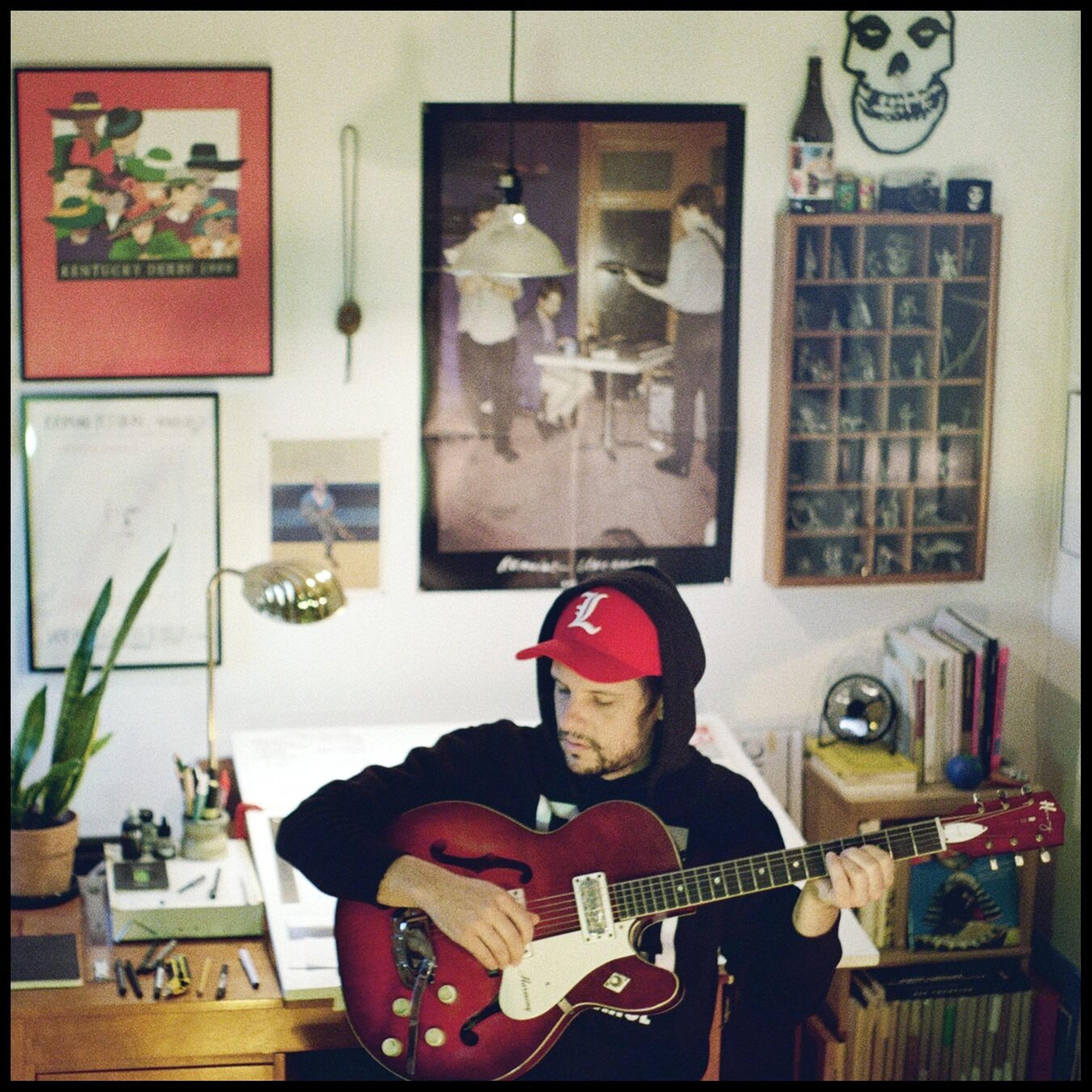Ryan Davis is sitting at a table playing guitar. He’s wearing a black hoodie and a red cap.