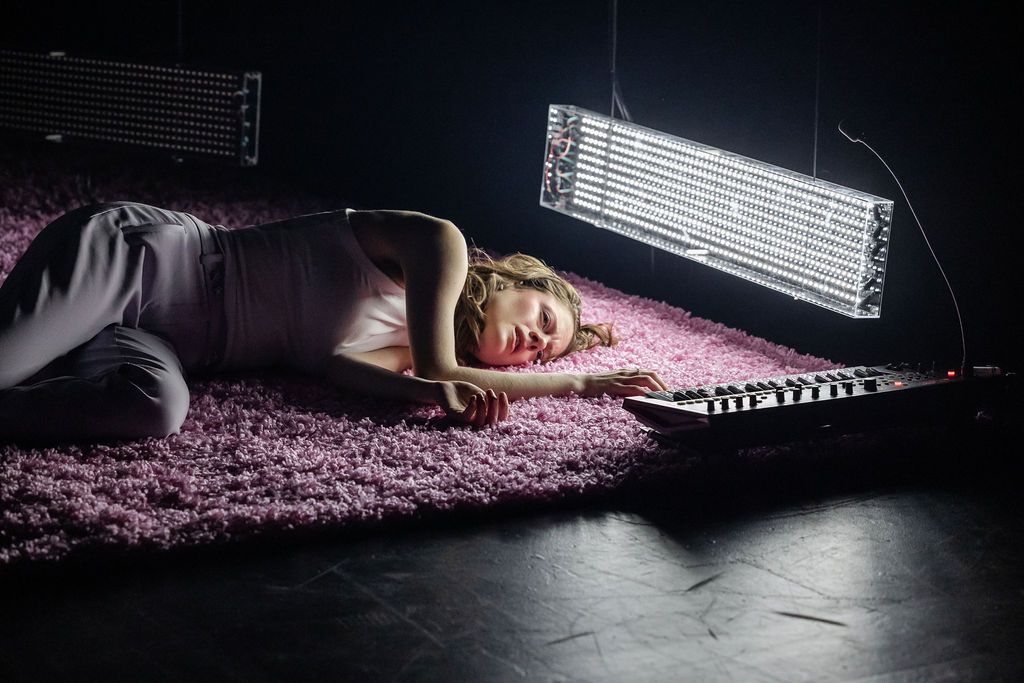Karin McCracken lies on carpeted floor with a florfloor light shining on their face and a keyboard on the floor 