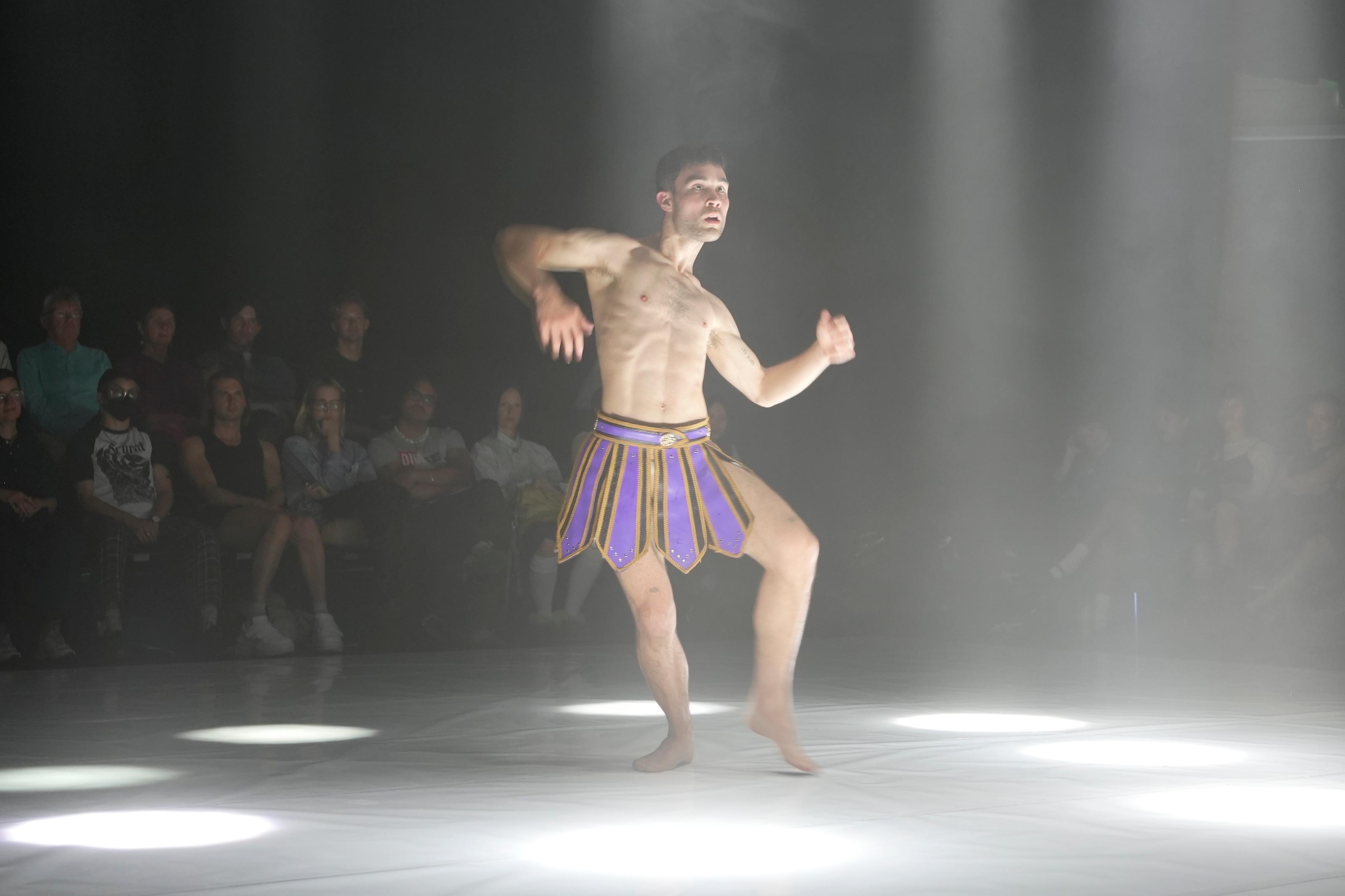 A performer wearing a skirt mid-choreography surrounded by spotlights and seated audience in the background.