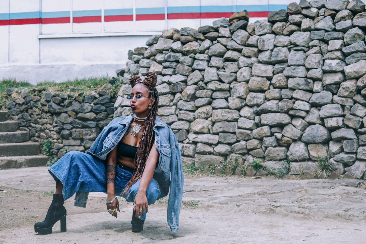 A person wearing denim and sunglasses squats in front of a stone wall, with long braided hair and a confident posture.