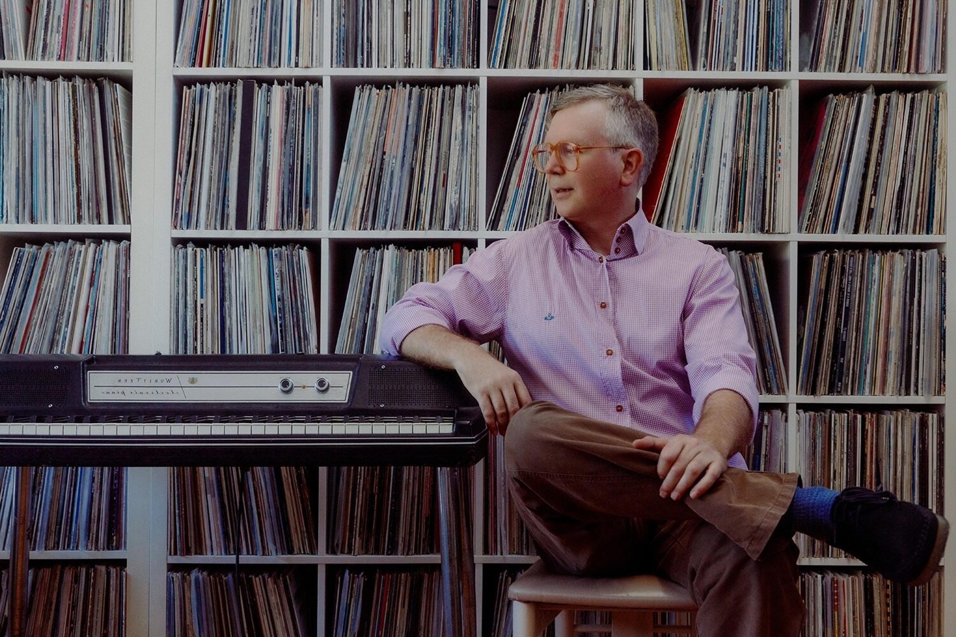 Guy Blackman is wearing glasses, sitting on a chair with his arm resting on an organ. He is looking sideways, crossing his legs, with a bookshelf in the background.