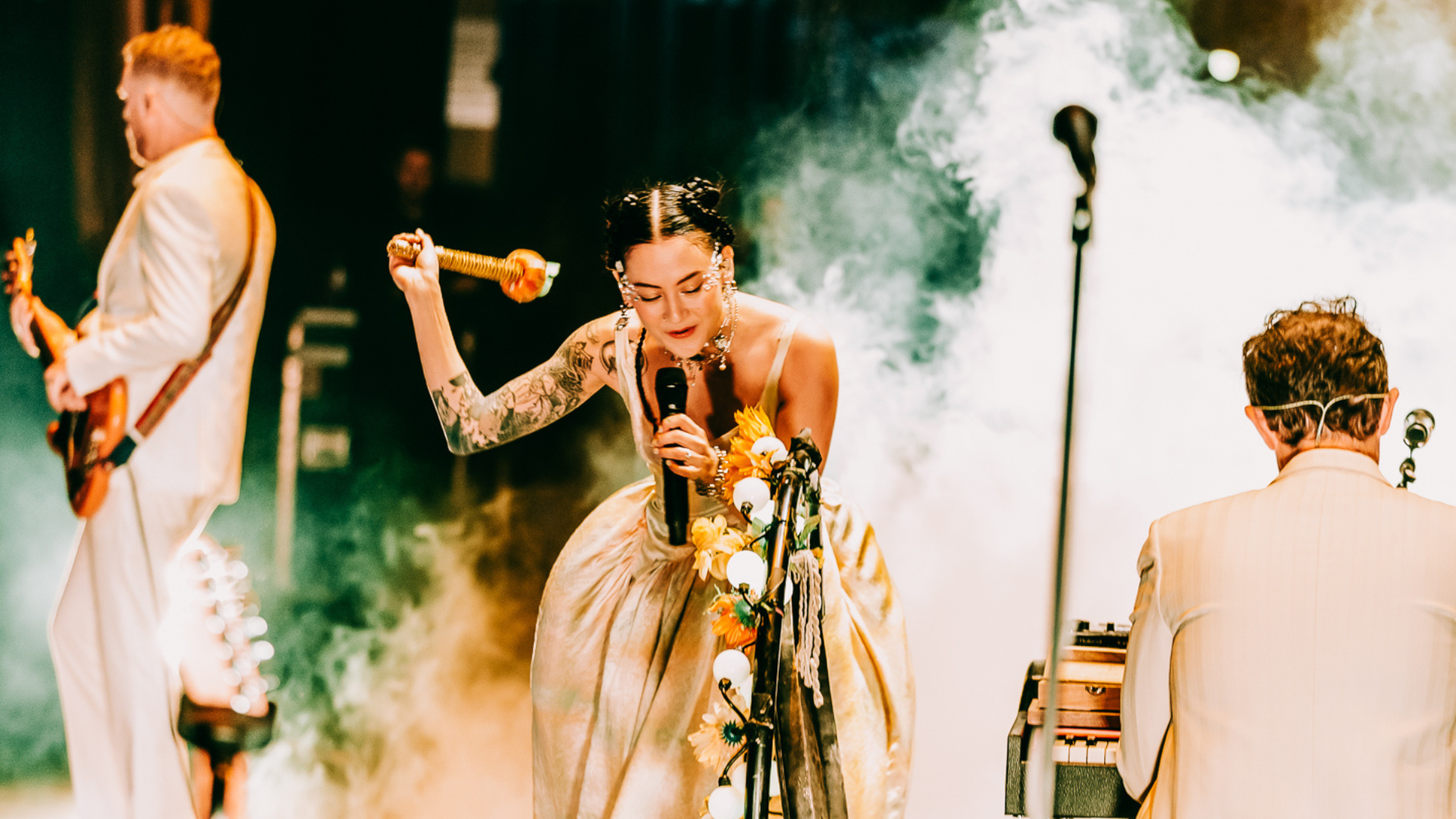 Japanese Breakfast performing on stage singing into a microphone and banging a gong, with band members in the foreground and background. 