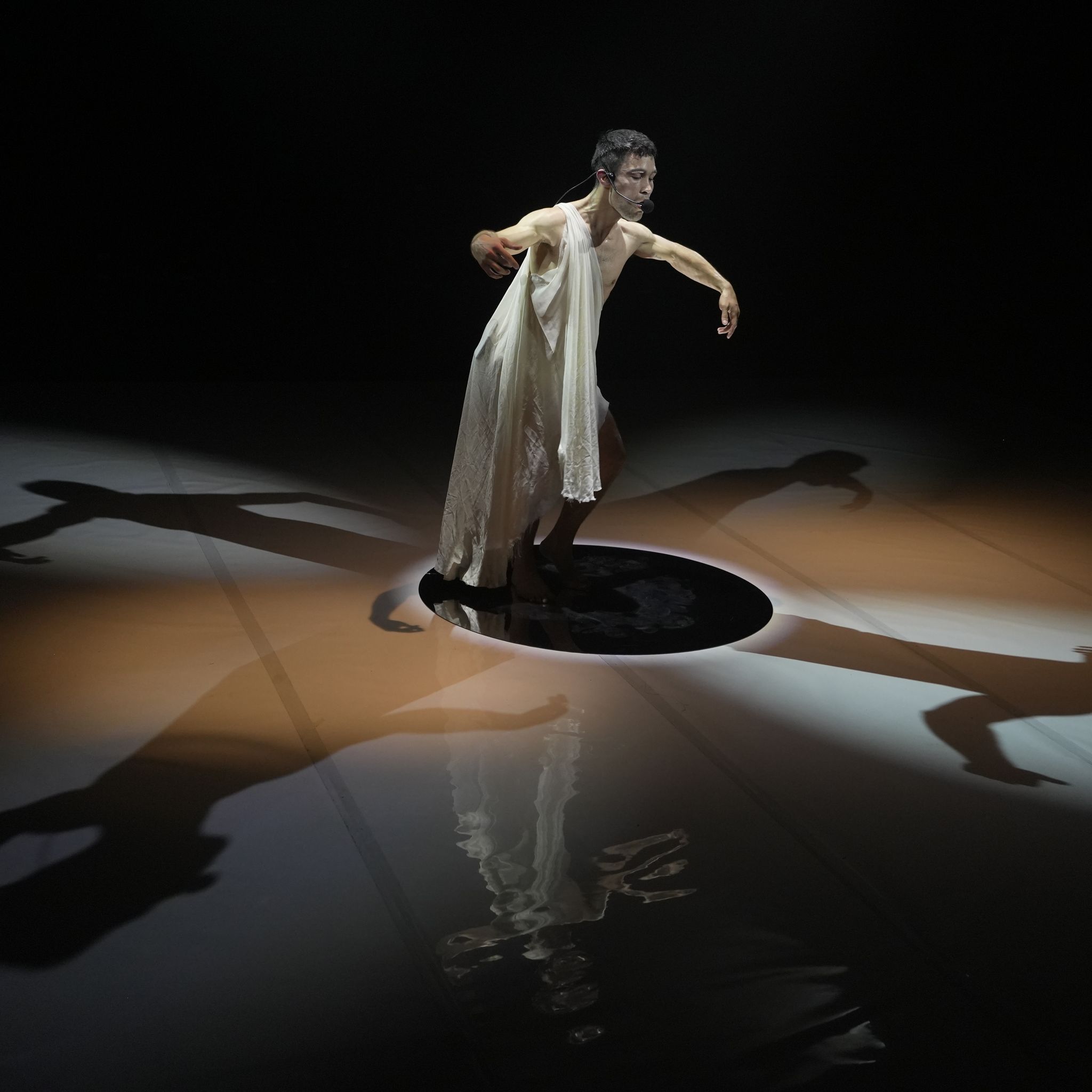 A performer wearing a flowy outfit posing dramatically on a dimly lit stage with their shadow cast from four different angles.