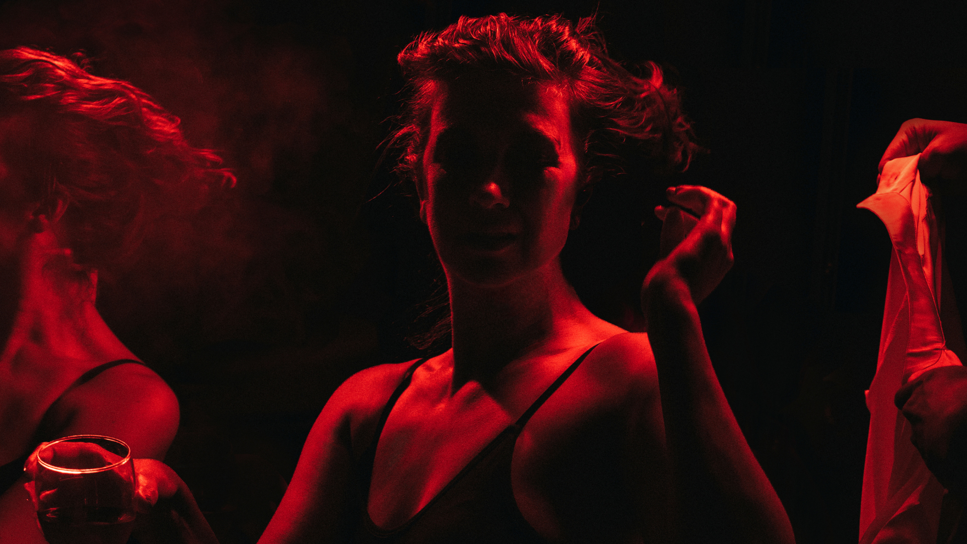 A dancer in a dress moves expressively, while a hand offers them a glass of wine and blurred figures shift in the background.