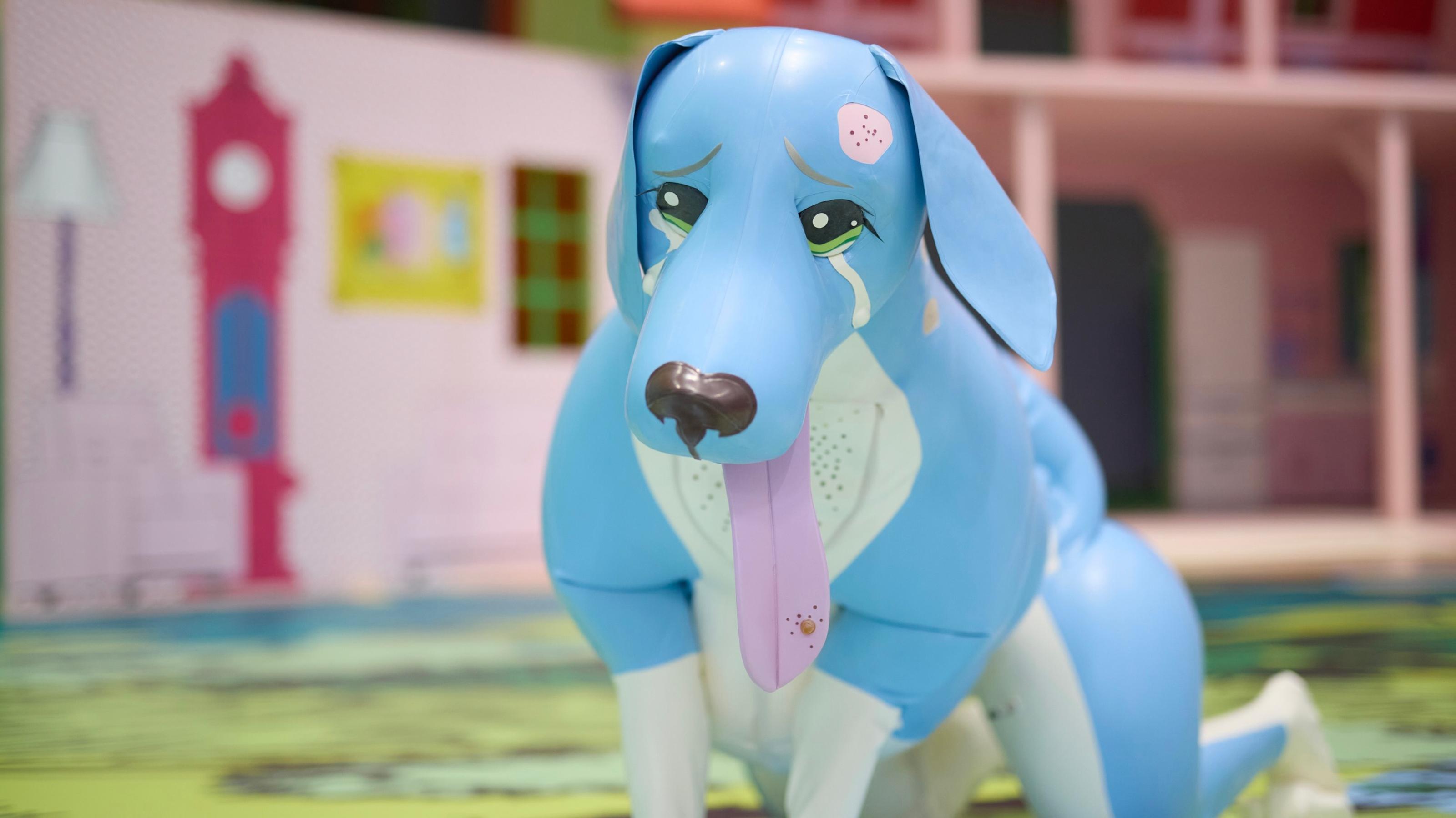 Large blue inflatable dog gazes sadly towards viewer, with tears streaming down face and its tongue sticking out.
