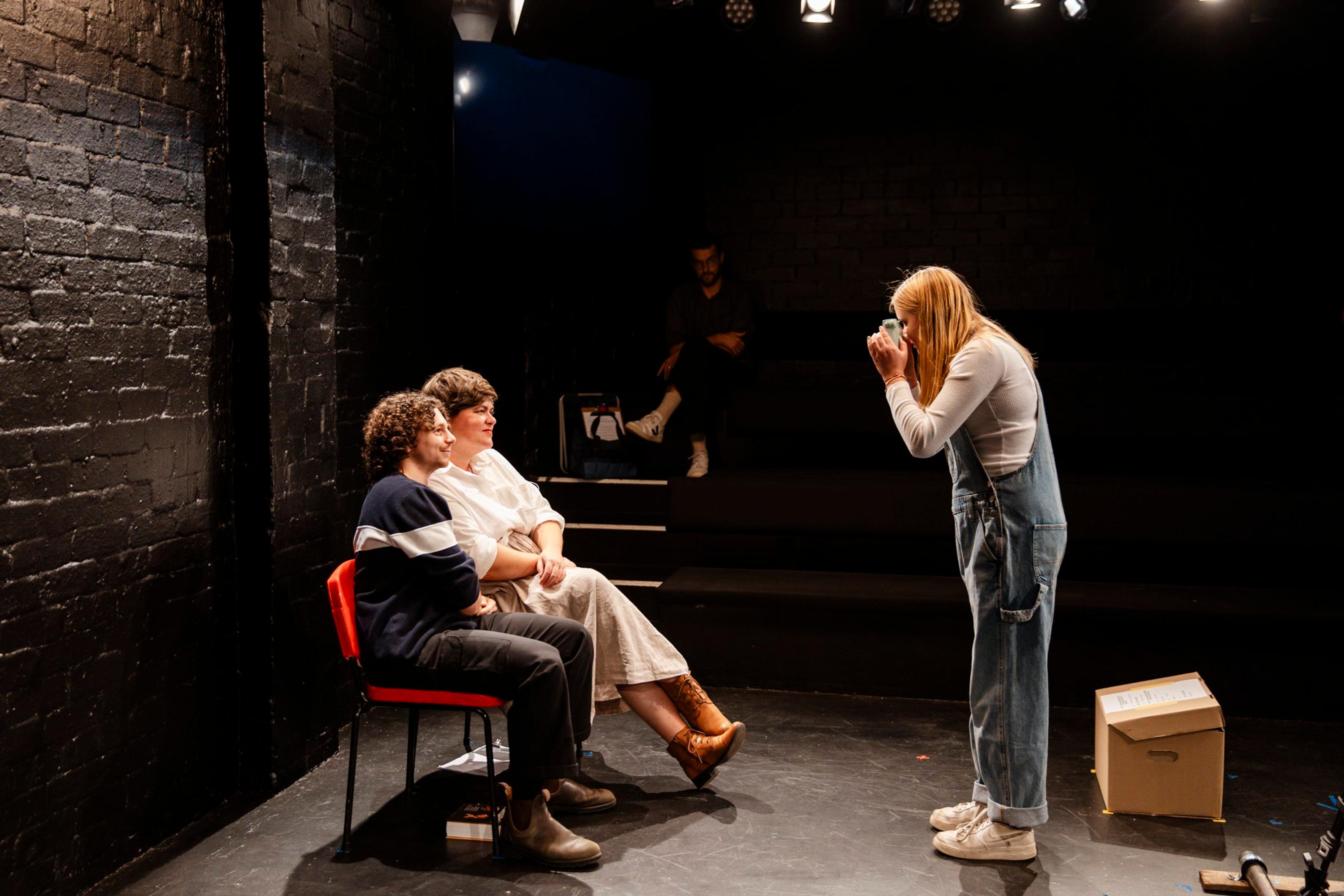 A young person holding a video camera films two adults seated together in a theatre space
